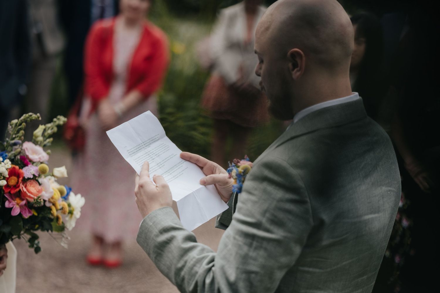 Person in grey suit reads from papers during outdoor wedding ceremony.