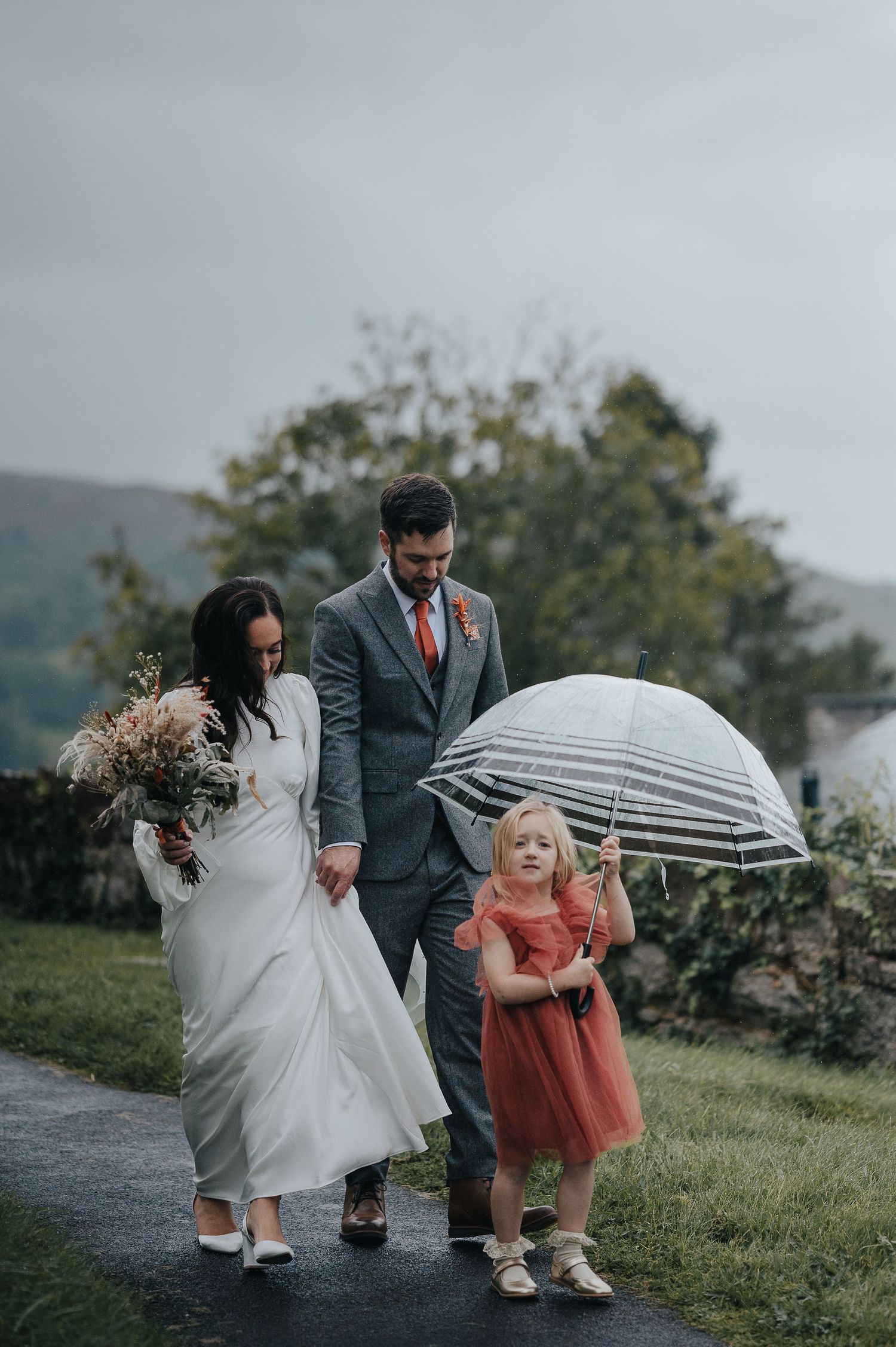 Wedding party walking in the rain with a clear umbrella as a young attendant in coral dress joins them.
