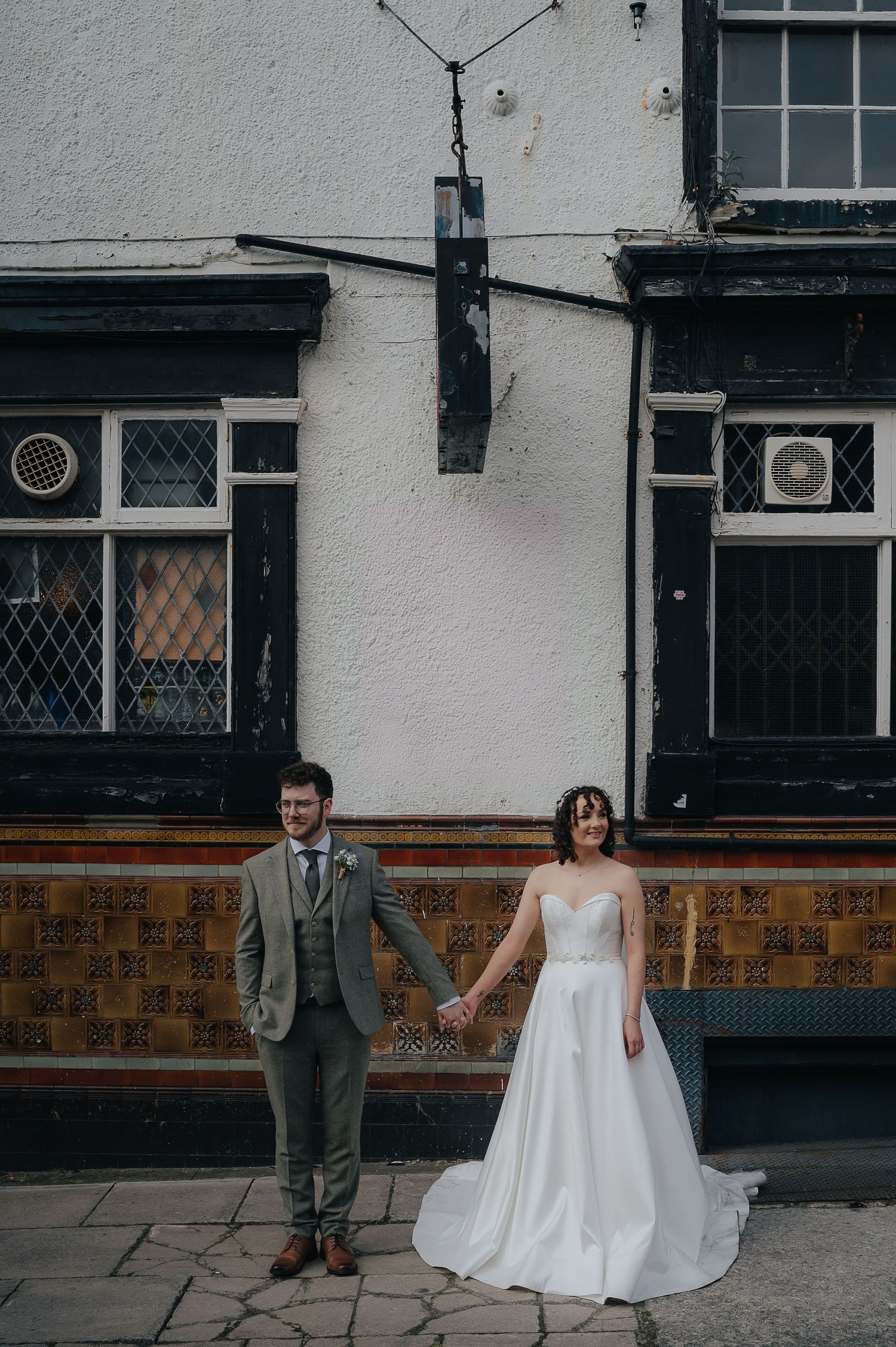 A couple posing in wedding attire against a white building facade with black trim.