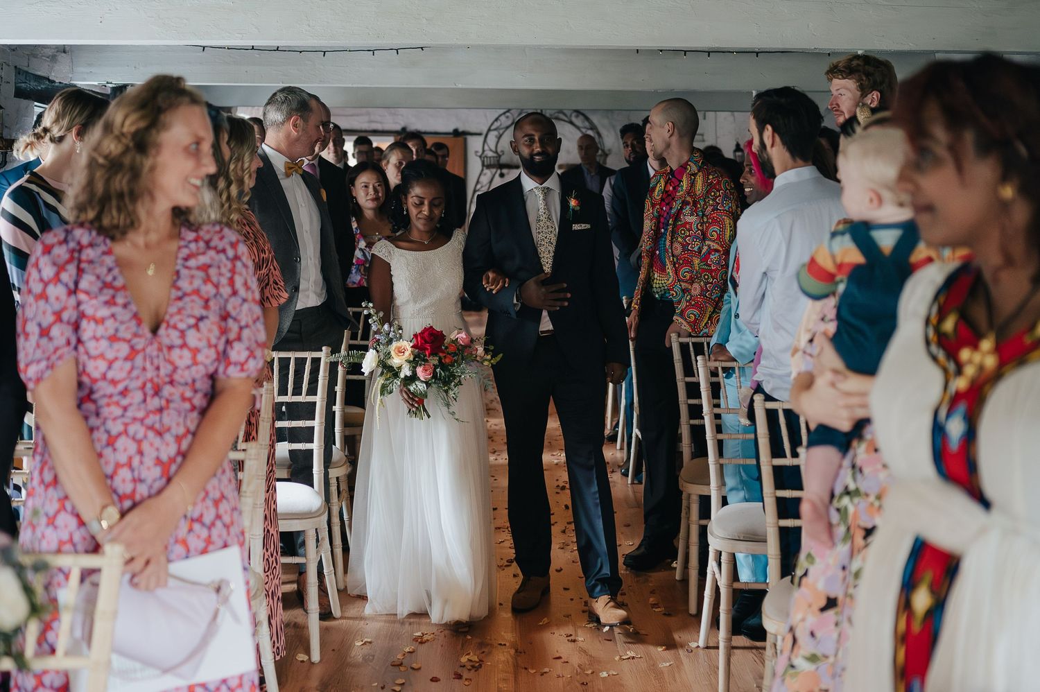 Wedding guests gather in rows of white chairs during an indoor ceremony.