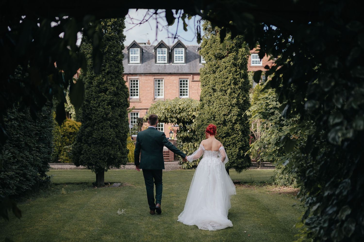 Wedding couple walks towards historic building through manicured garden path.