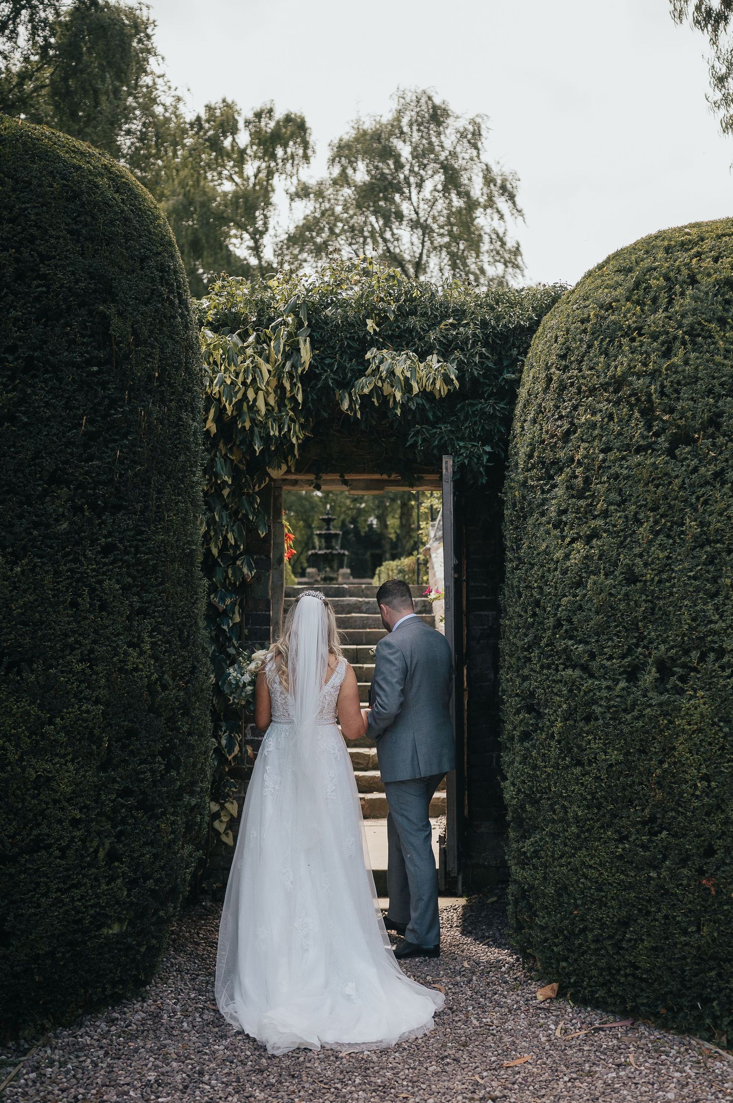 Wedding couple walking through manicured hedge archway leading to garden steps.