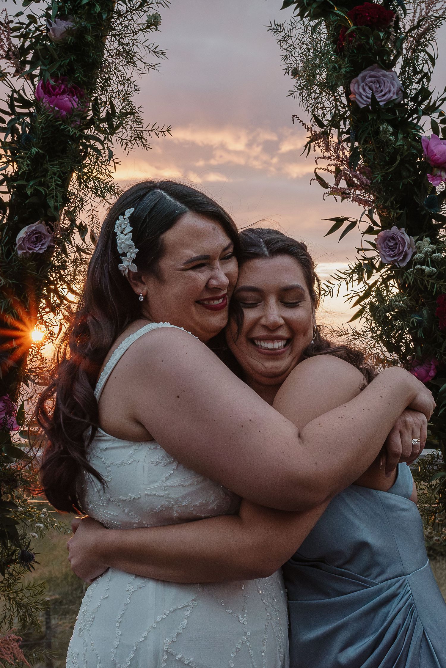 Two people embracing under a floral arch during sunset.