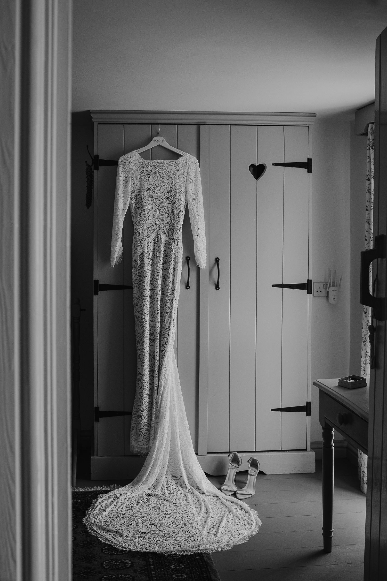Elegant lace wedding dress with long train hanging against wooden paneling in black and white.