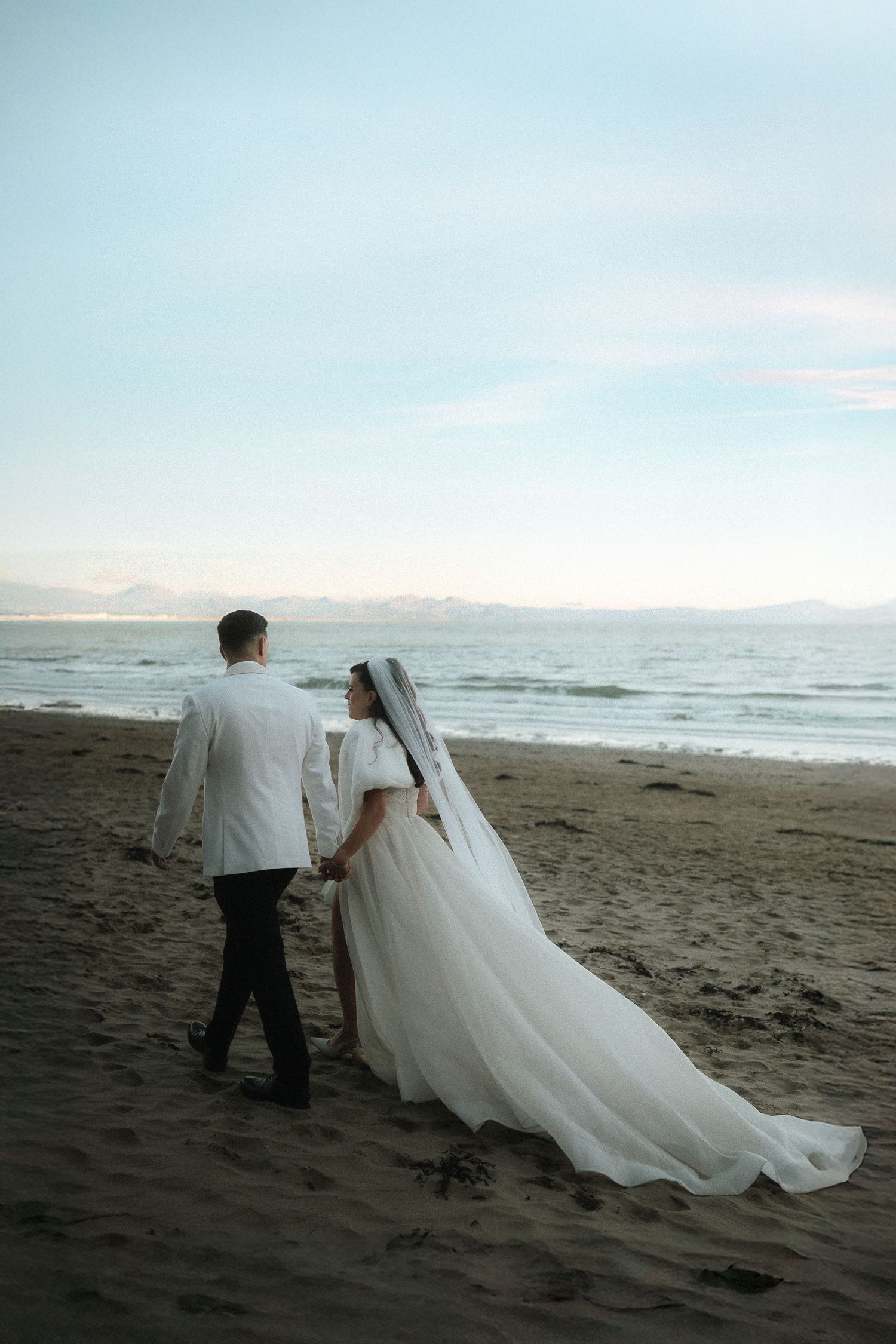 A couple in wedding attire walks along a misty beach at sunset, with the bride's white dress flowing in the wind.