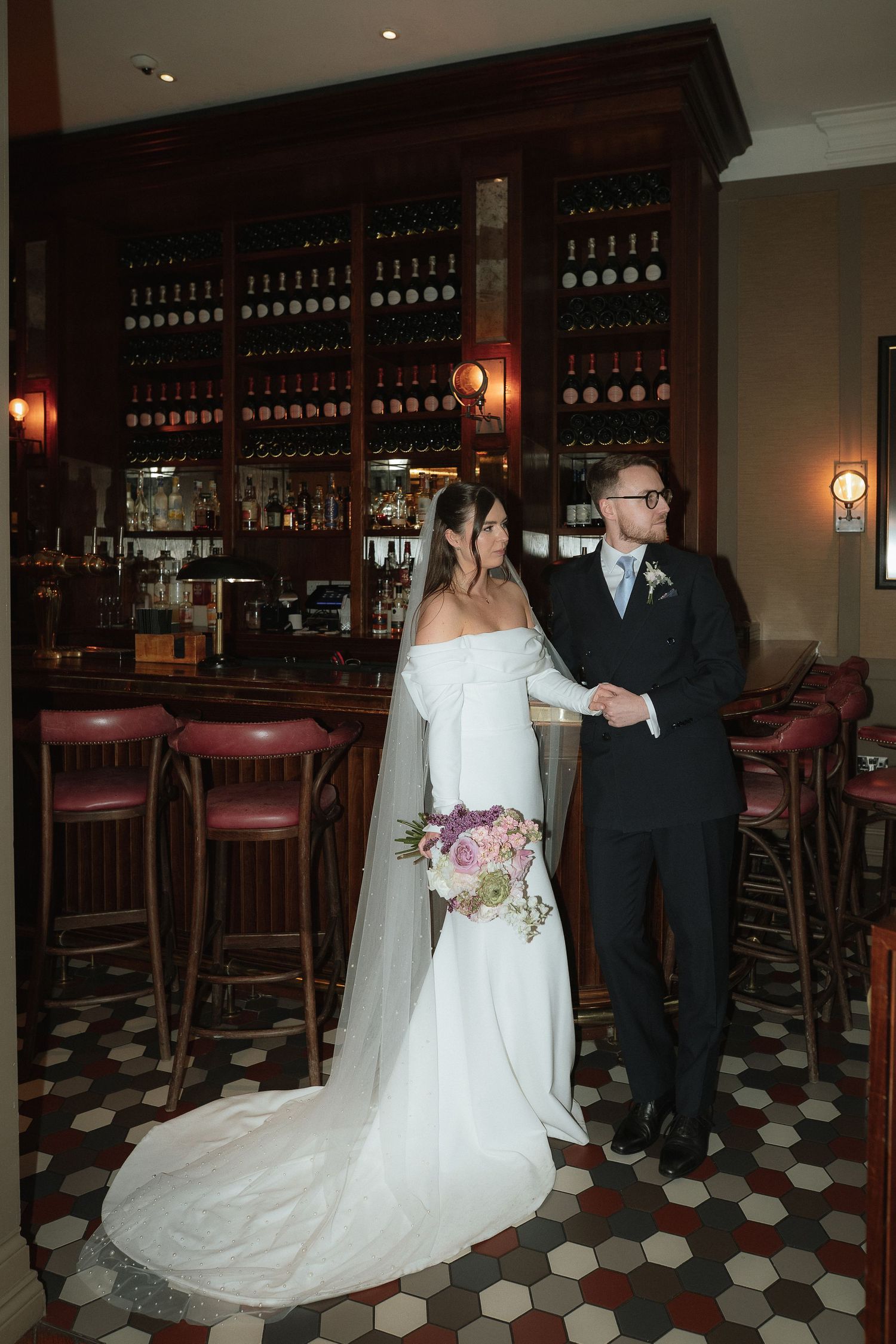 Bride and groom pose elegantly at a classic bar venue, bride in off-shoulder gown holding purple bouquet.