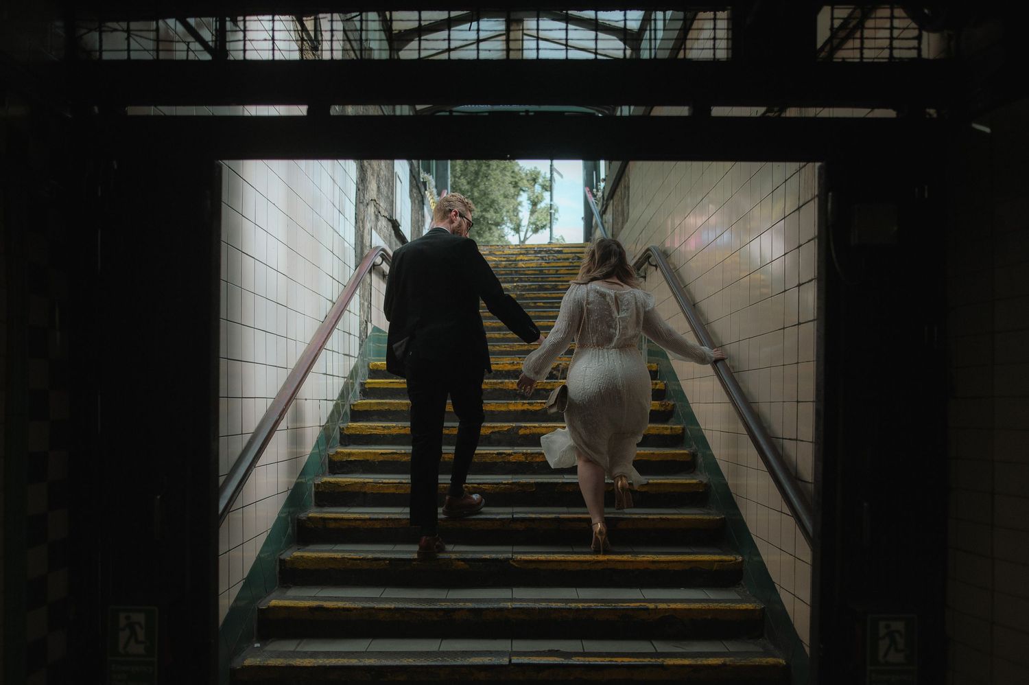 A dark subway station corridor with silhouetted figures walking up and down stairs in a repeating pattern.