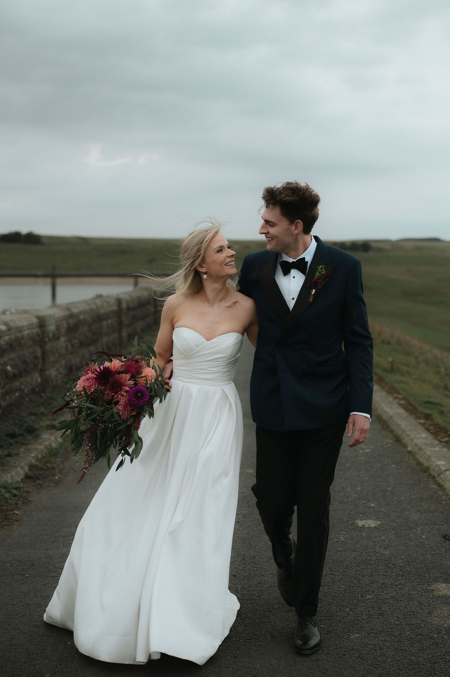 A couple walks together on a countryside path, the bride holding a vibrant burgundy bouquet. Blackton Grange Wedding