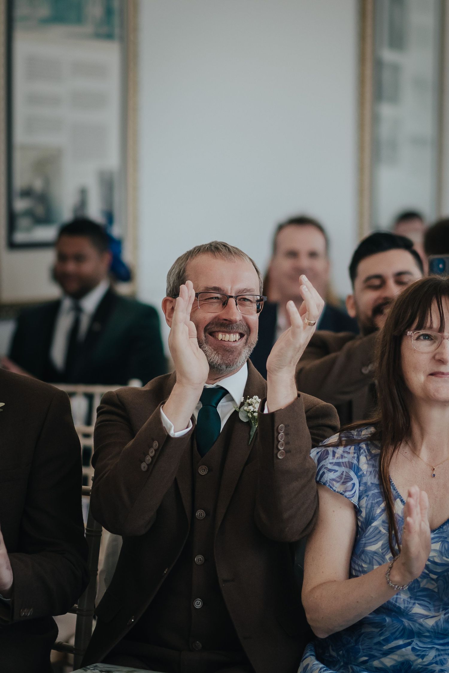 A guest reacting with joy during a wedding ceremony while wearing a brown suit.