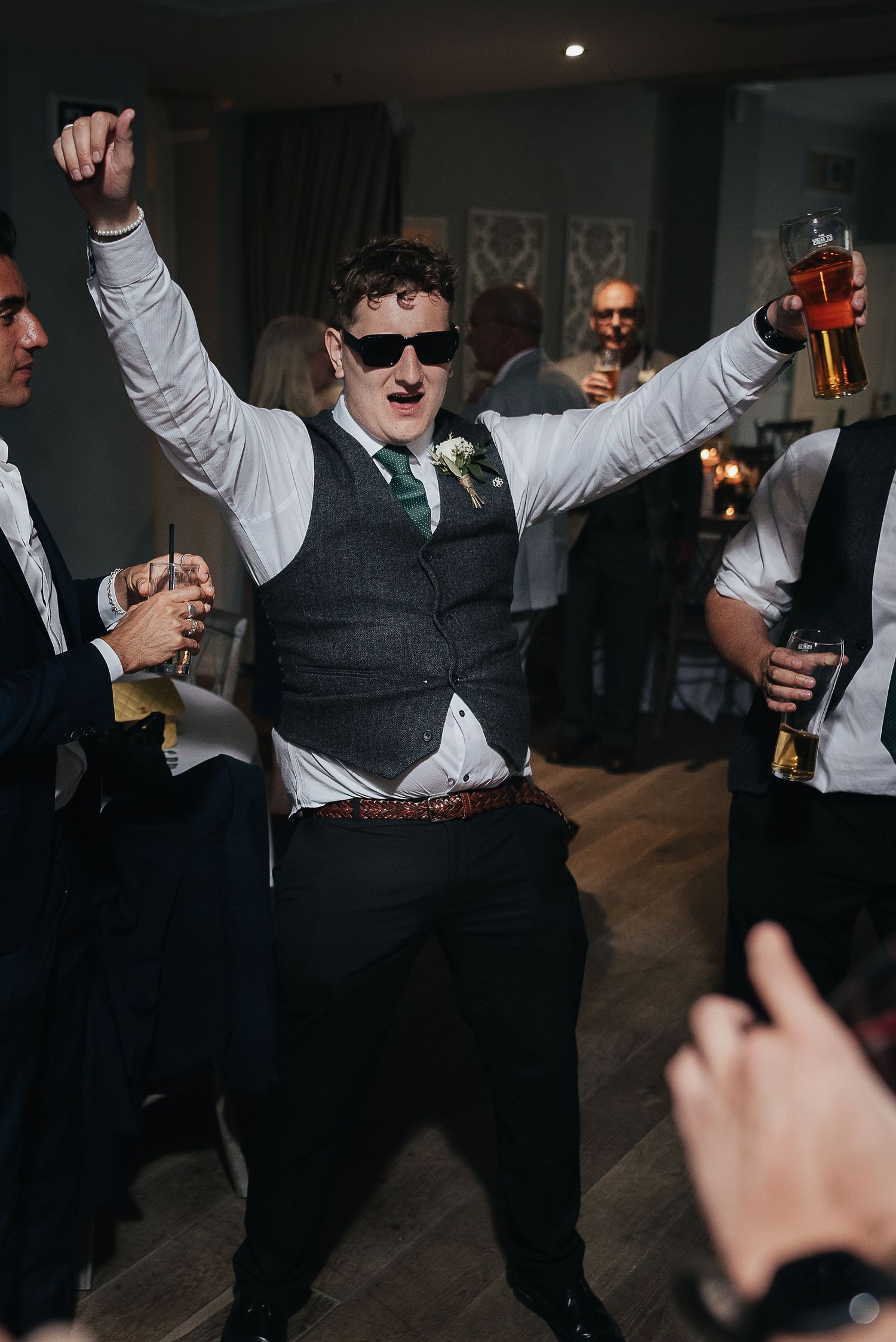 Party guest in waistcoat and sunglasses enthusiastically dances while holding drinks in both hands.