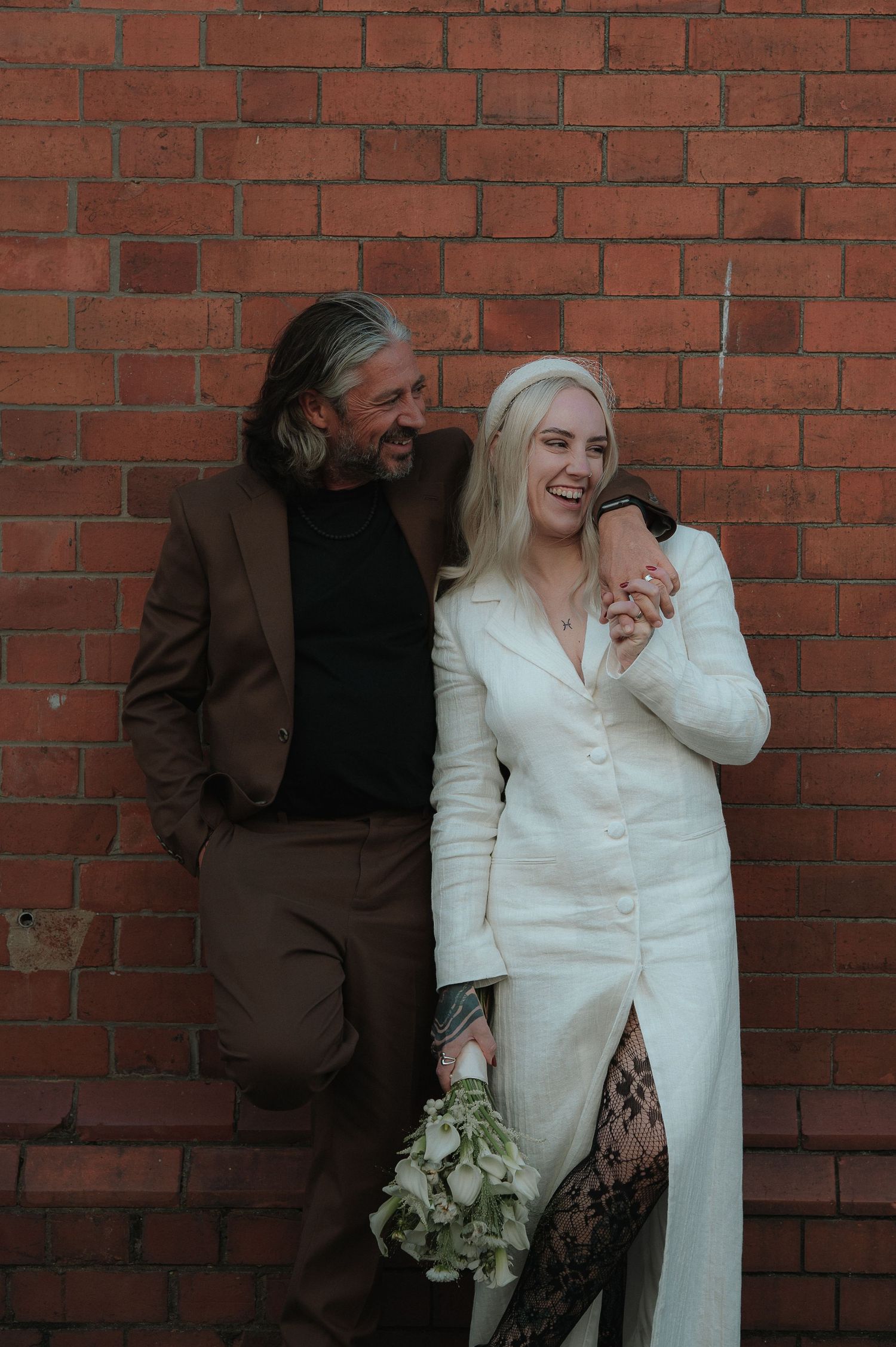 Wedding couple posing against a red brick wall with white attire and floral bouquet.