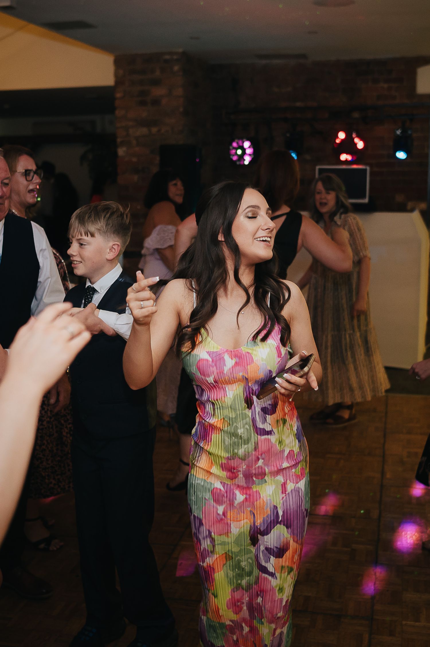 Guest dancing in colorful floral dress at wedding reception with brick wall backdrop.