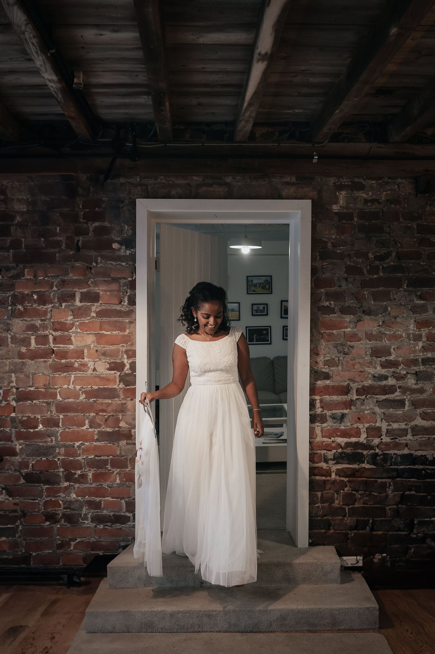 A bride in a flowing white dress stands in a doorway framed by exposed brick walls.