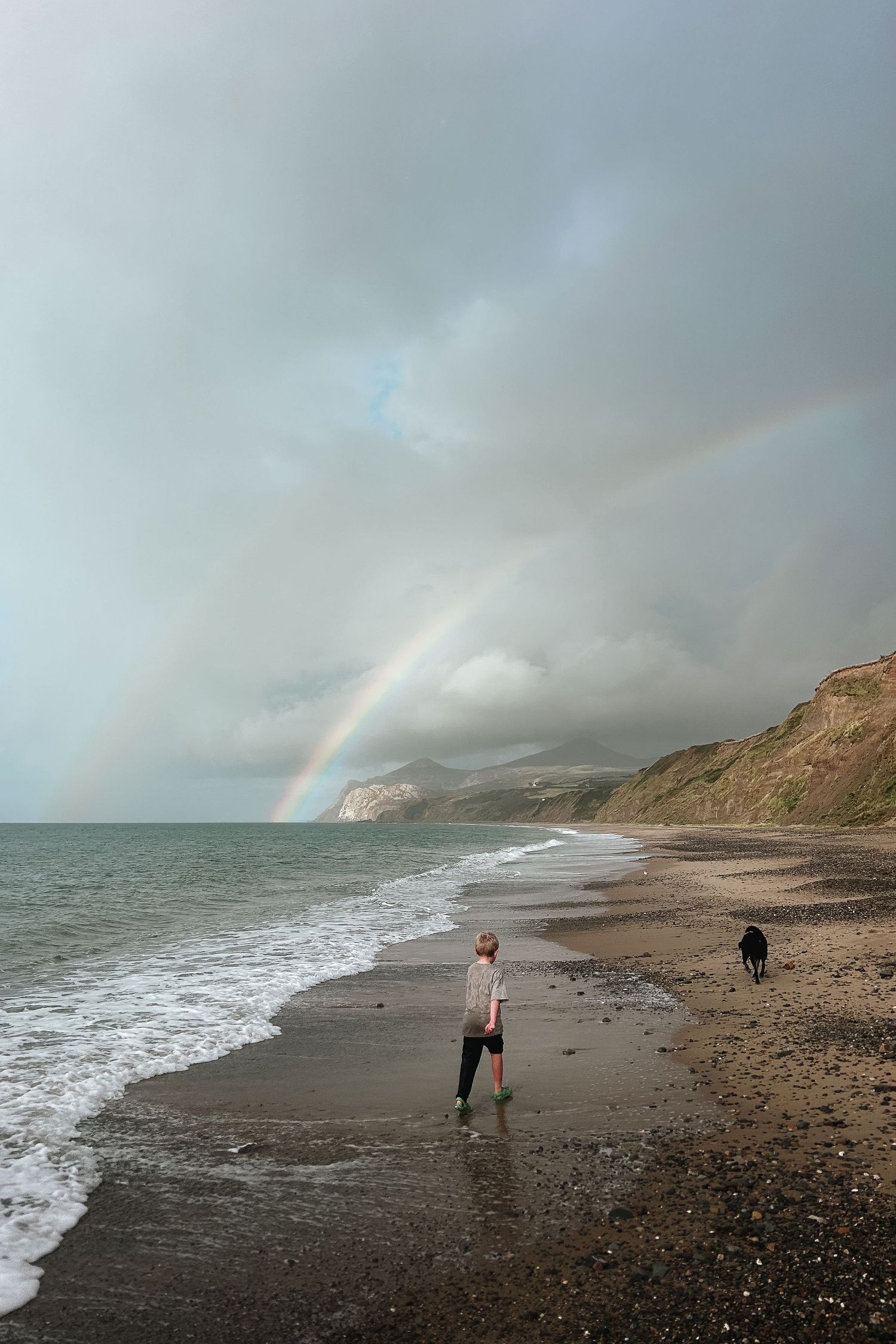 A rainbow arches over a dramatic coastal landscape with figures walking along a rocky beach beneath stormy skies.