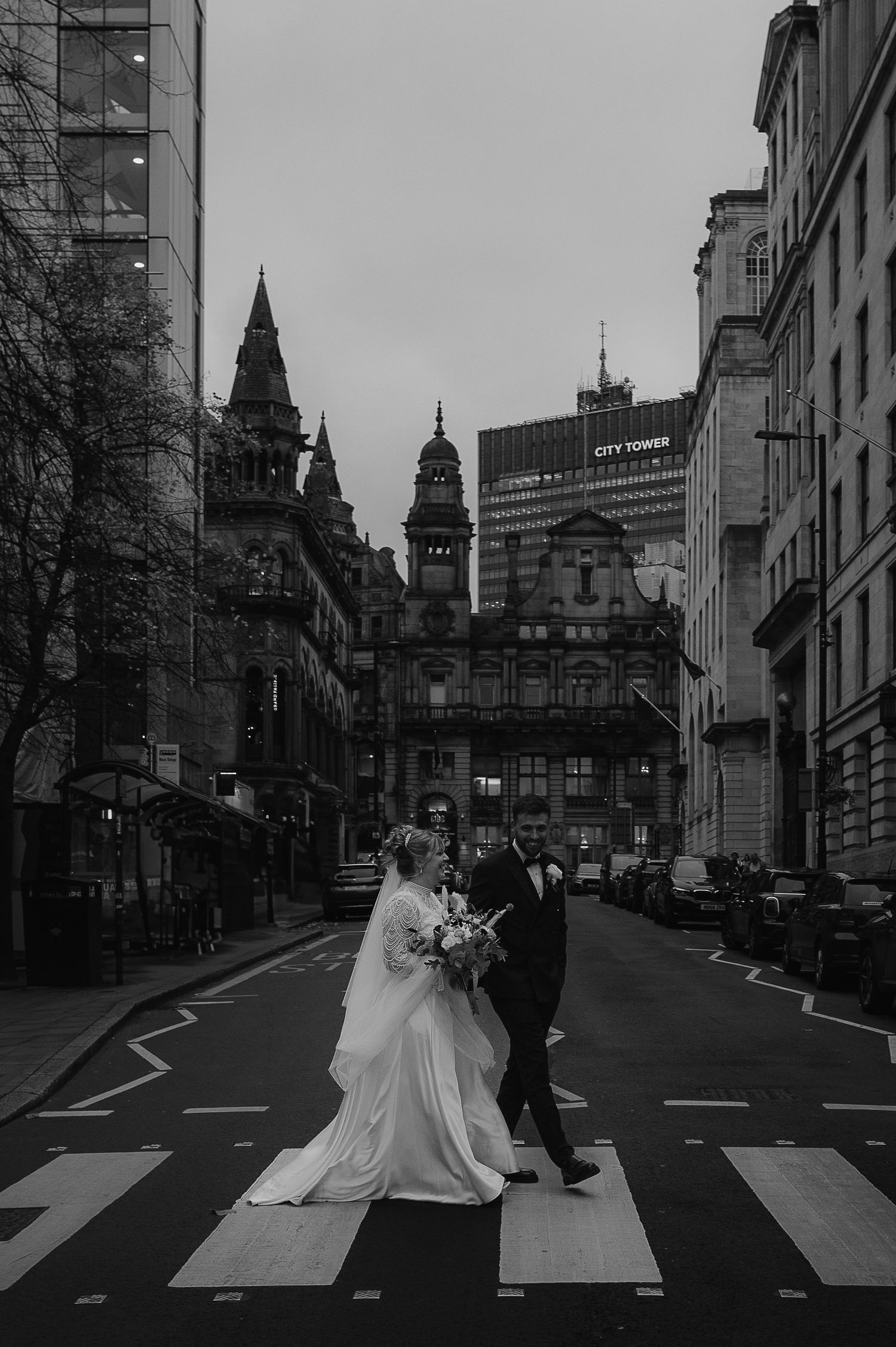 A couple in wedding attire walks across a crosswalk in a city street with gothic architecture in the background.