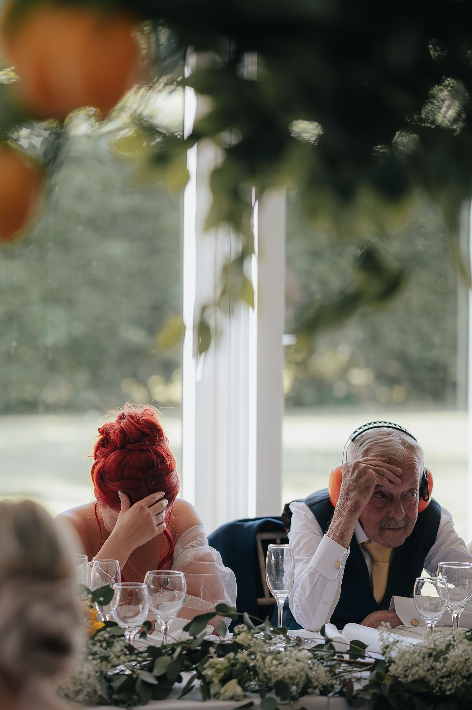 Wedding guests at reception table with one covering face in an emotional or humorous moment.
