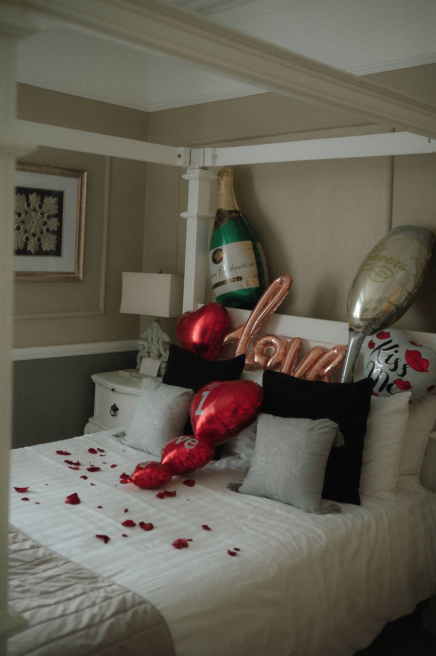 Romantic hotel room setup with white bedding decorated with scattered red rose petals and balloons.