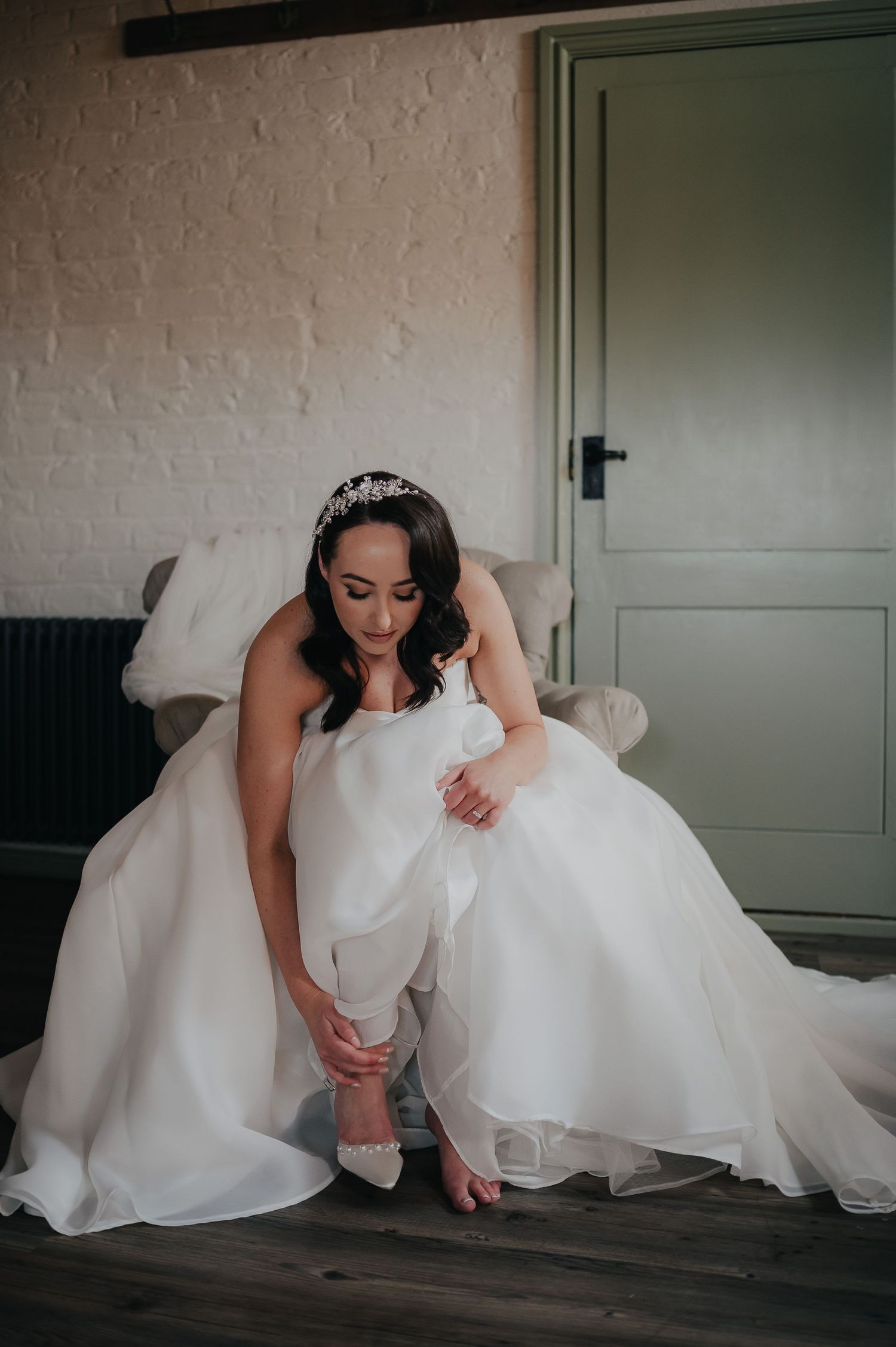 Bride in white gown adjusts her shoe while sitting in a minimal room setting.