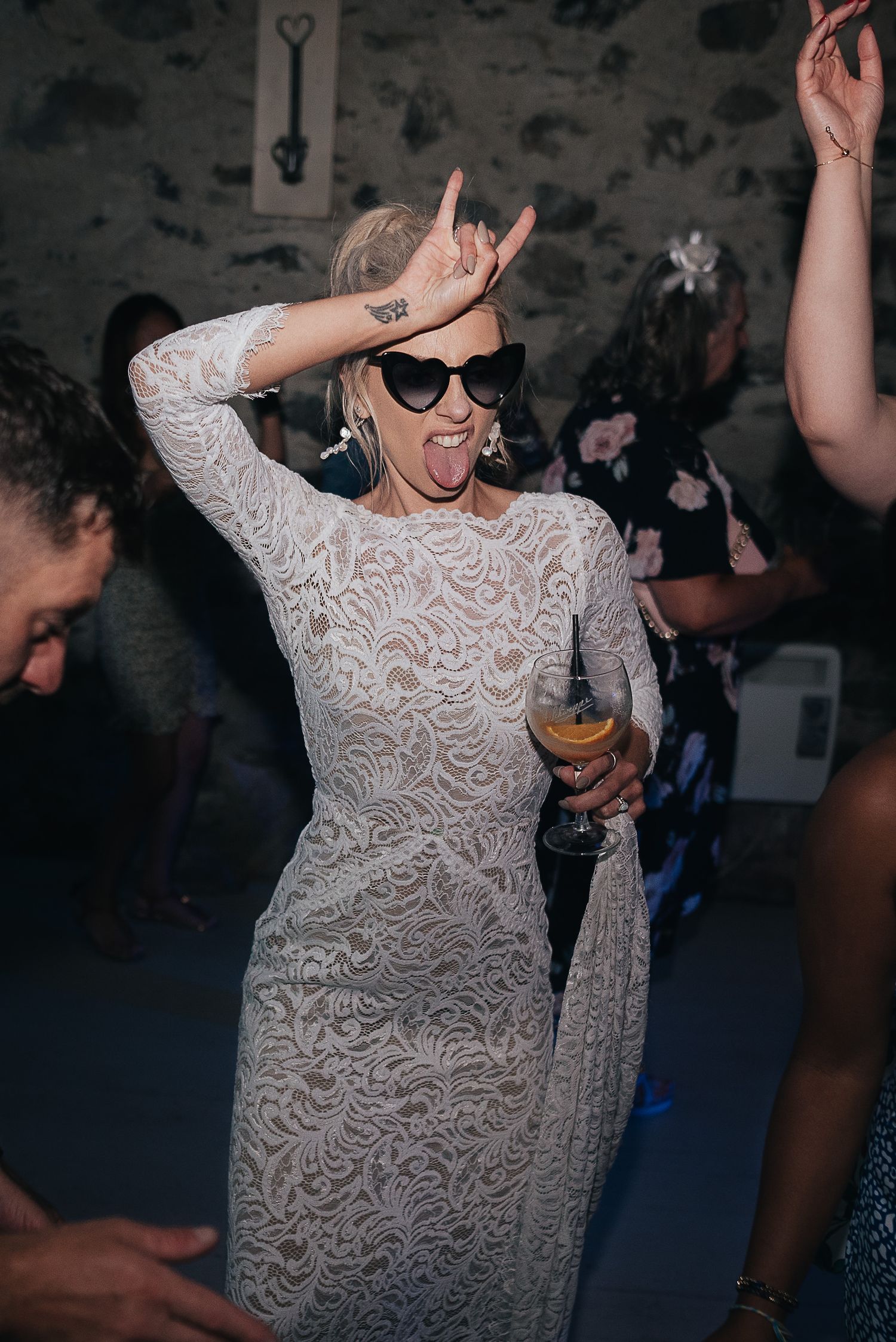 A person in a white lace dress dances energetically at a party while wearing sunglasses.