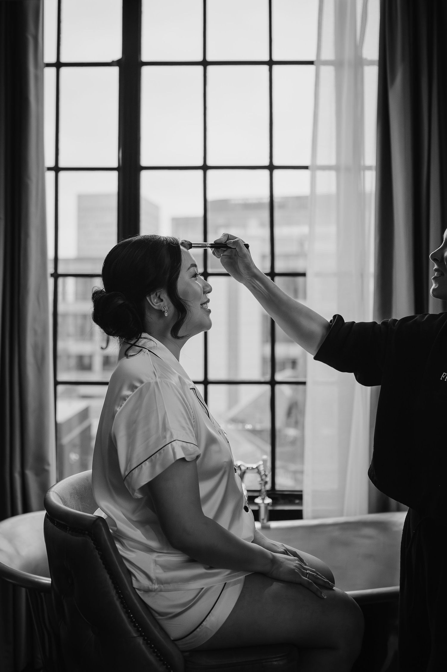 Black and white photo of bridal makeup preparation by window light.