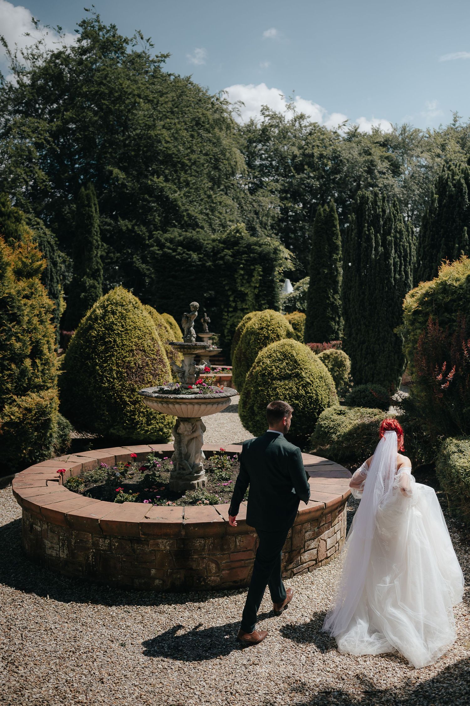 A wedding couple walks through a manicured garden with topiary bushes and a stone fountain.