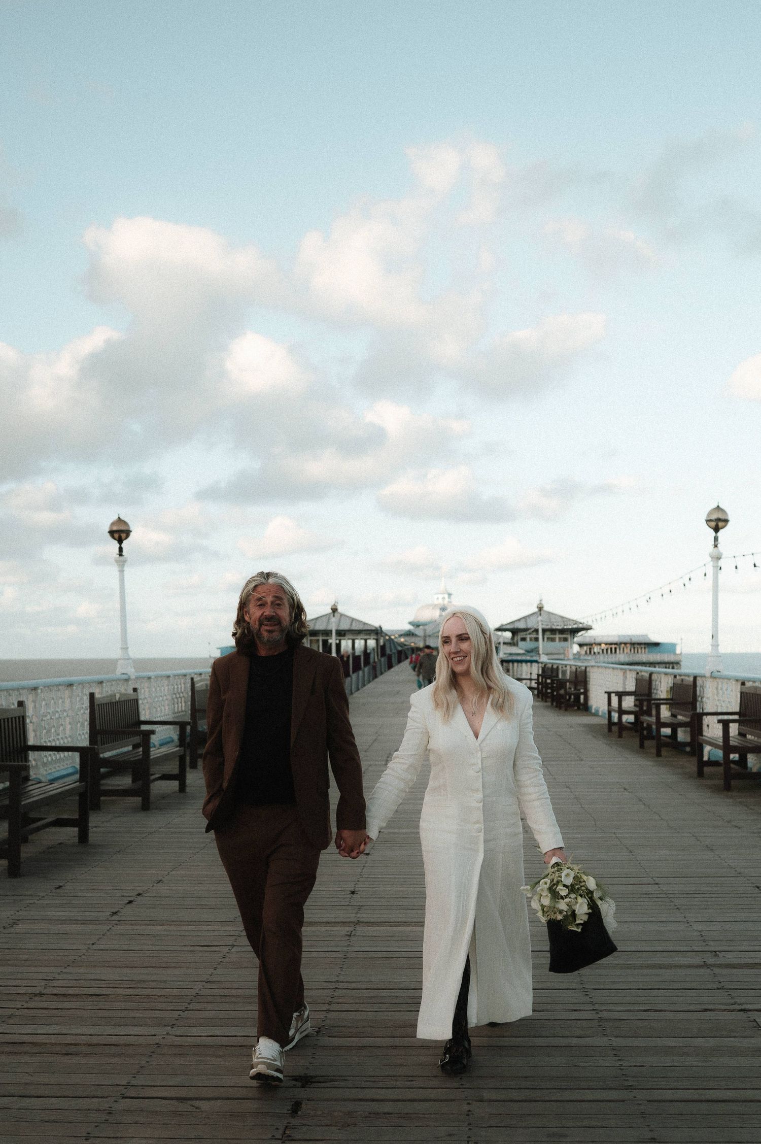 Couple walking hand in hand along seaside pier under cloudy sky.