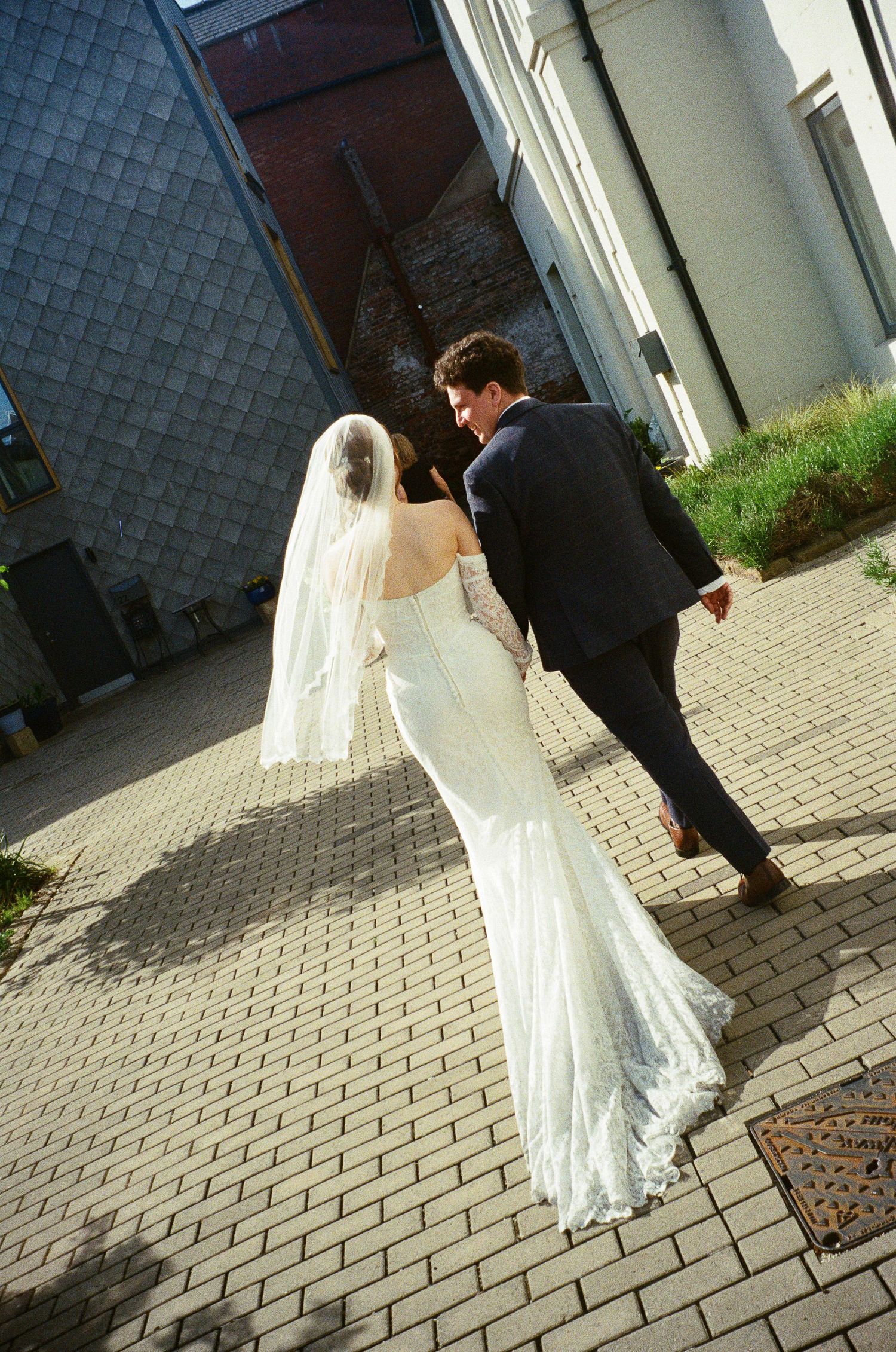 Bride in white lace gown and veil walks with groom in dark suit on cobblestone path near building.