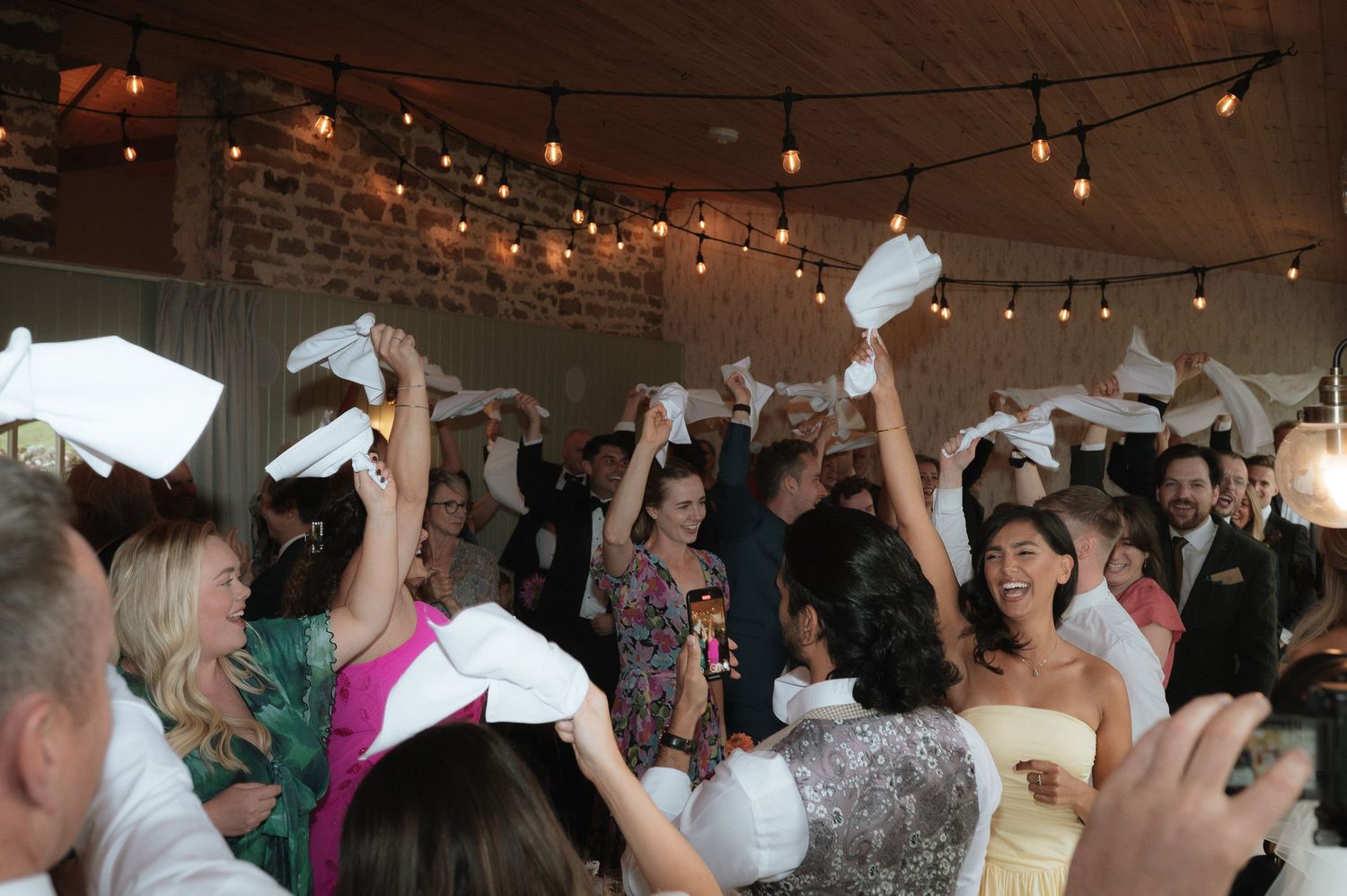 Wedding reception guests wave white napkins in celebration under string lights at an indoor venue.