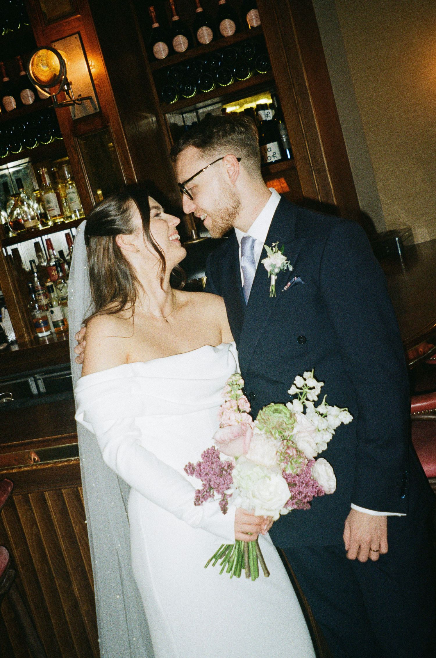 Bride and groom share a romantic moment, she holds a pink and white bouquet, inside an elegant bar venue.