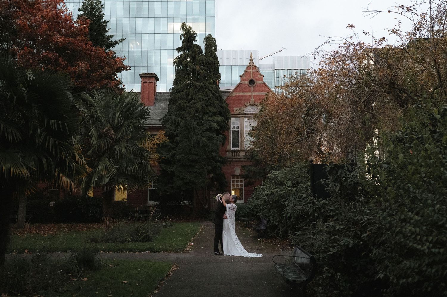 Bride and groom embracing in lush garden grounds before a historic red brick building surrounded by trees. Manchester Wedding Photographer.