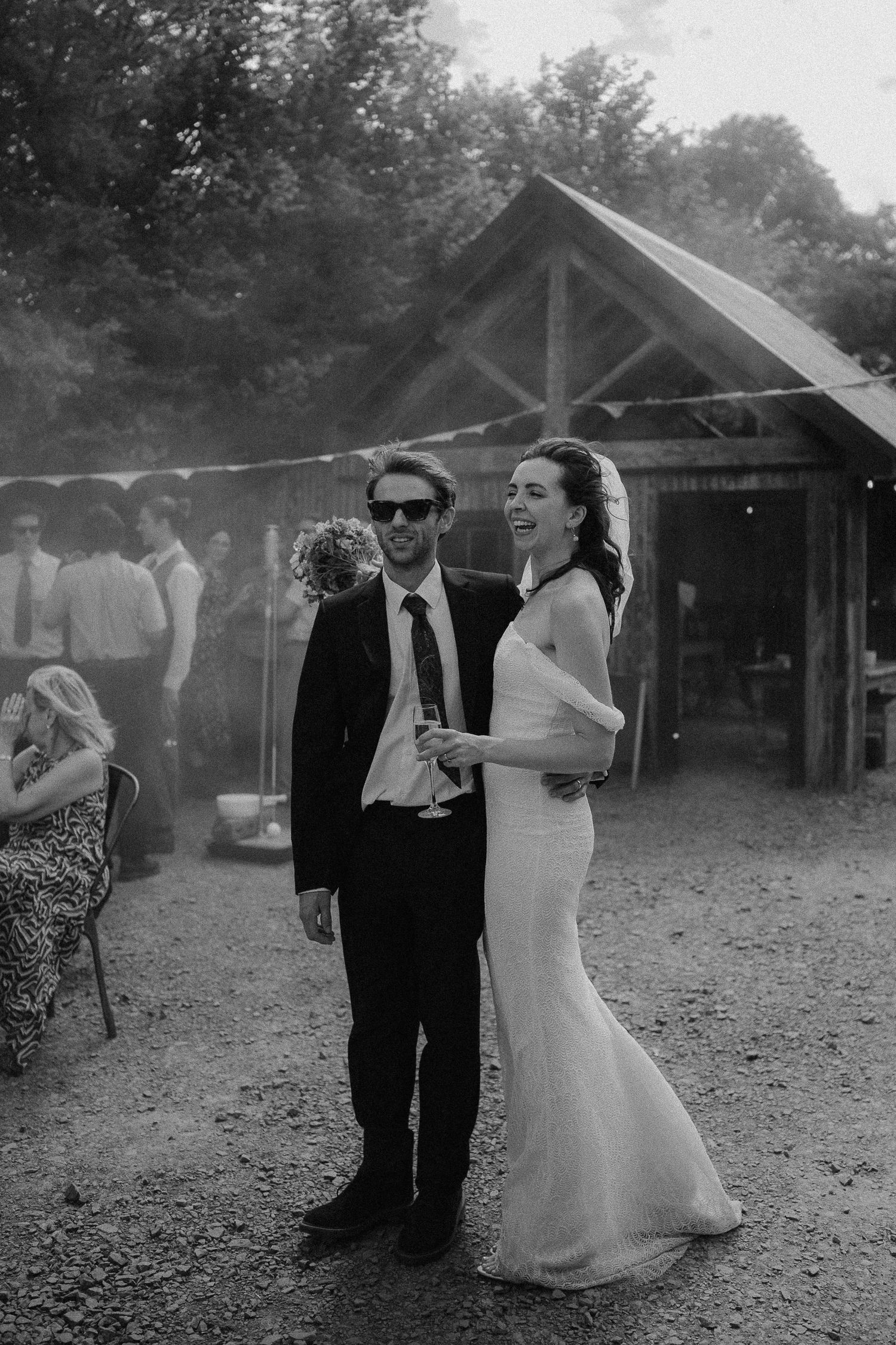 Black and white photo of a smiling bride in a fitted gown and groom in a suit with sunglasses at an outdoor wedding reception.