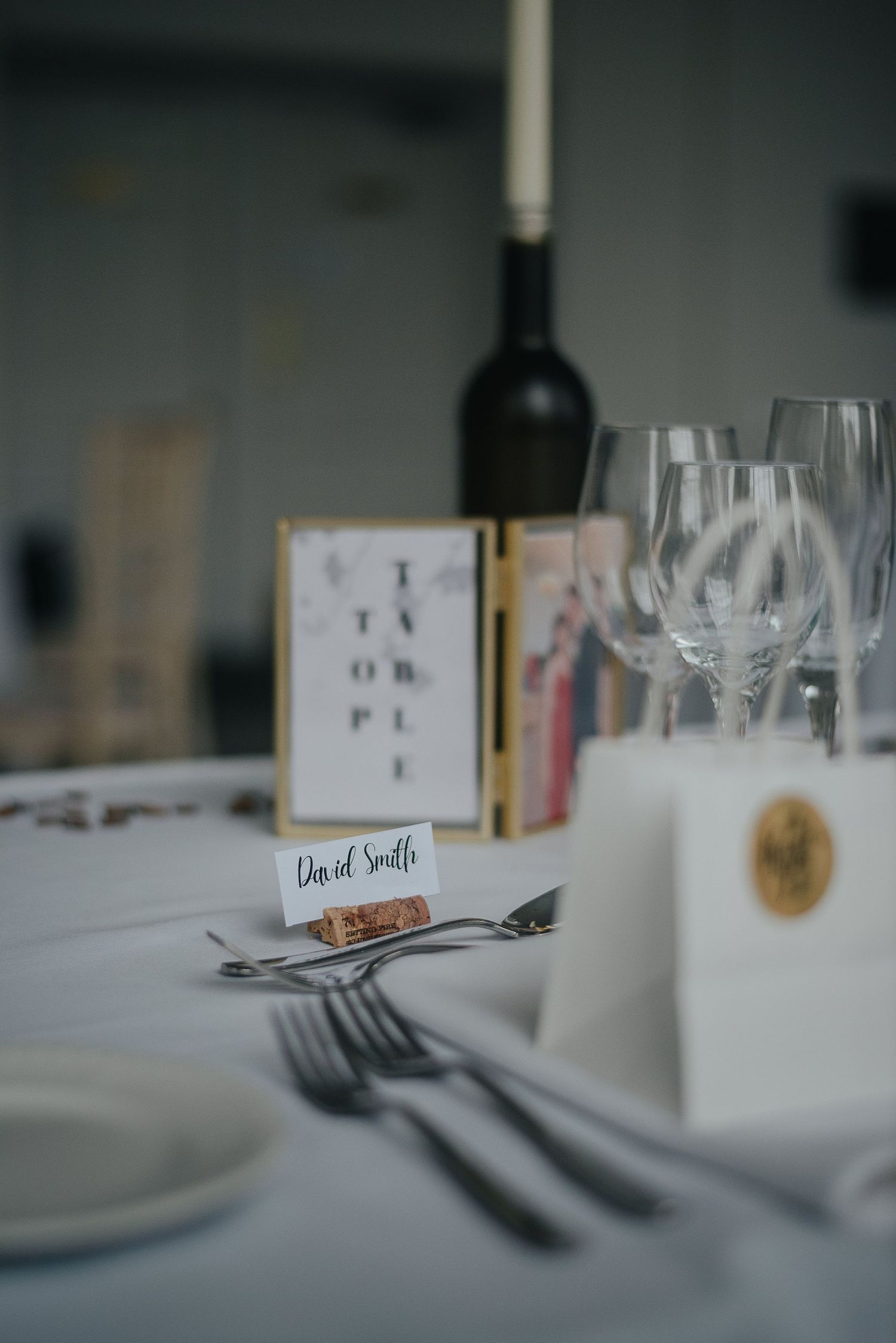 Place setting at wedding reception with wine bottle and name card on white tablecloth.