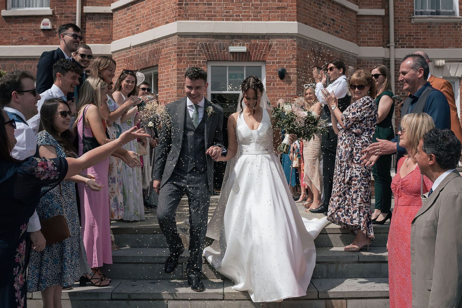 Wedding couple exiting venue while guests toss confetti on brick steps.