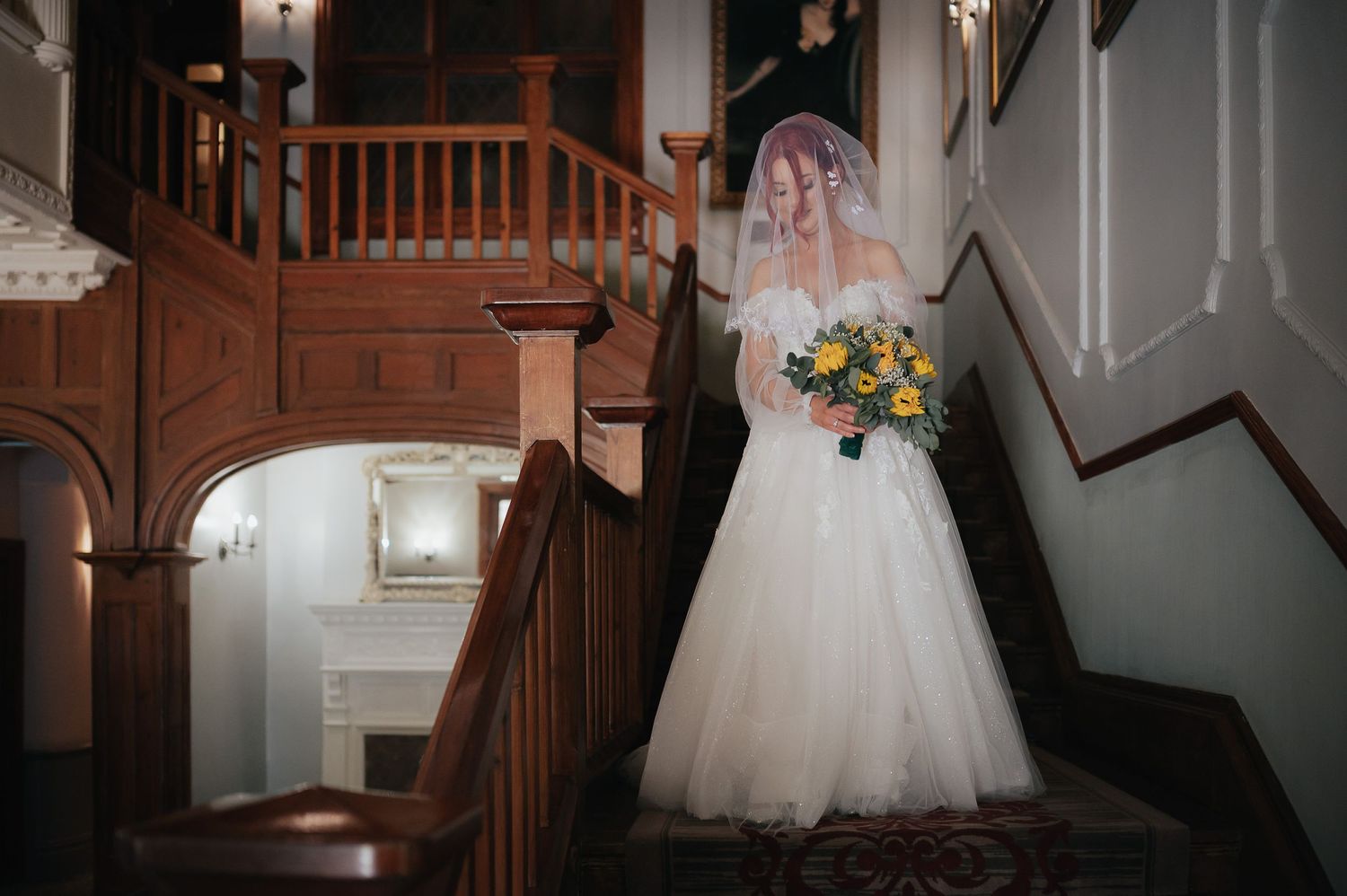 Bride in tulle wedding dress stands gracefully on vintage wooden staircase.