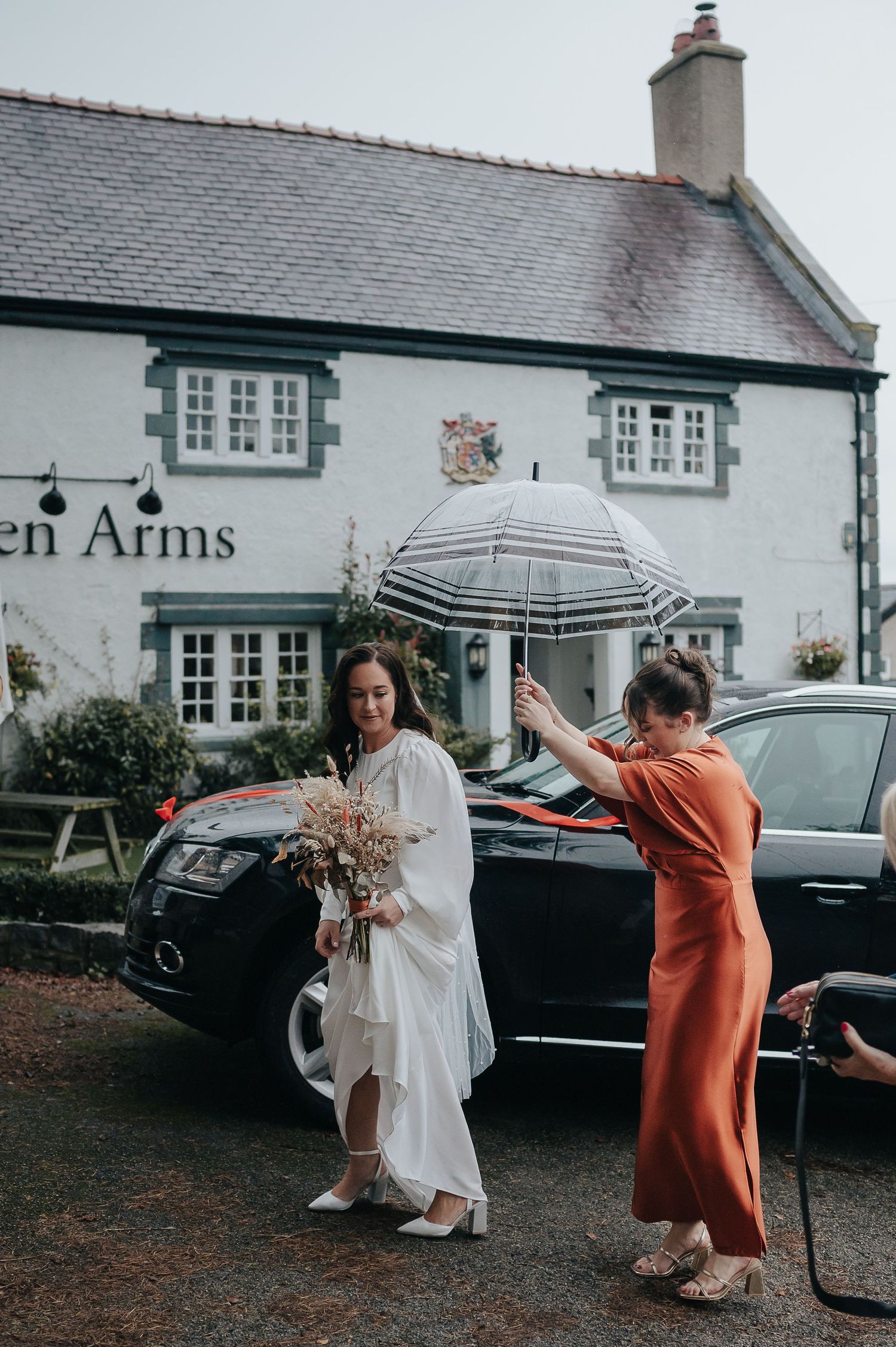 Outside a pub called Arms, a bride shelters under a striped umbrella on a rainy day.