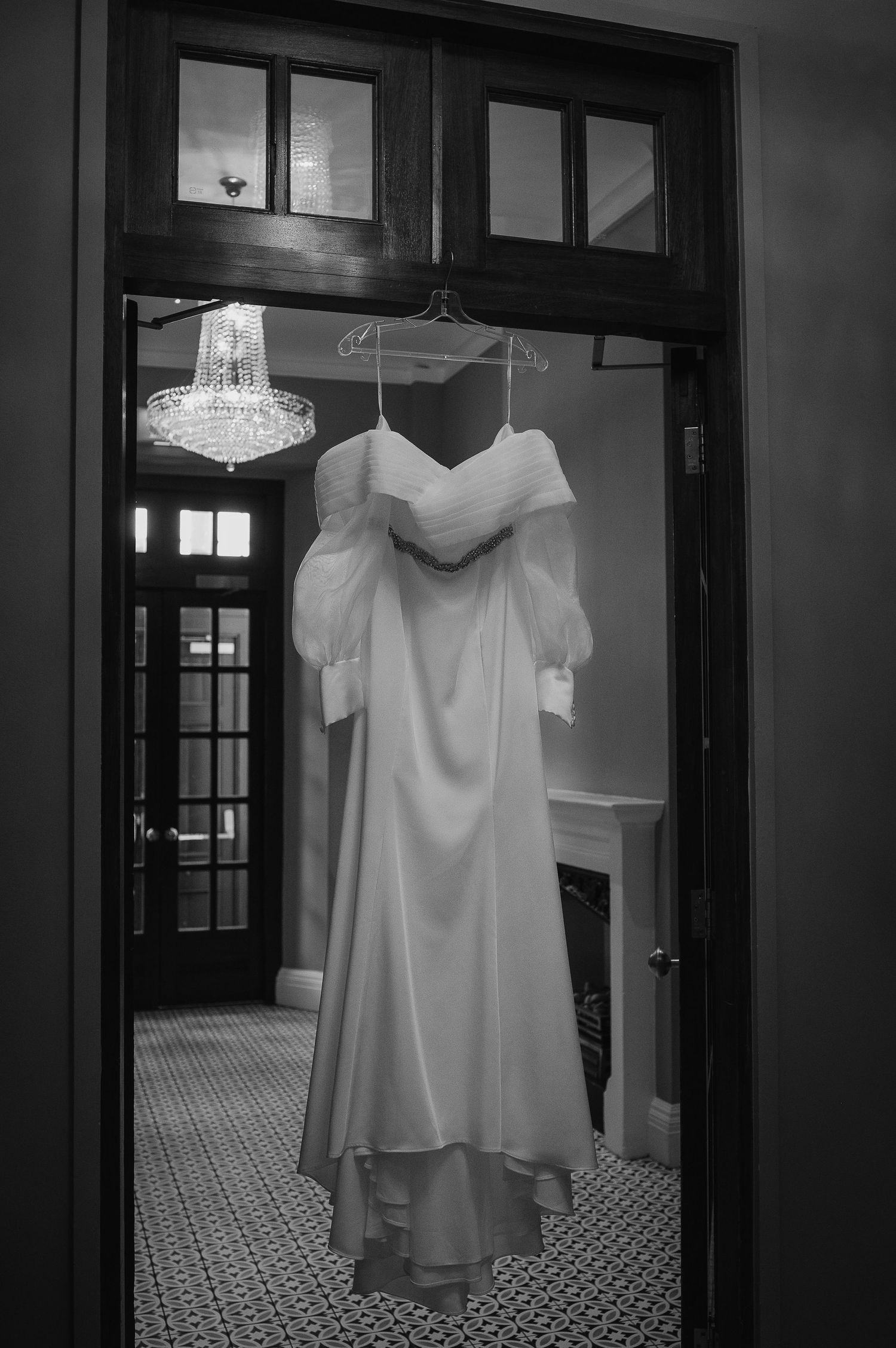 An elegant white wedding dress hangs in a doorway beneath a crystal chandelier.