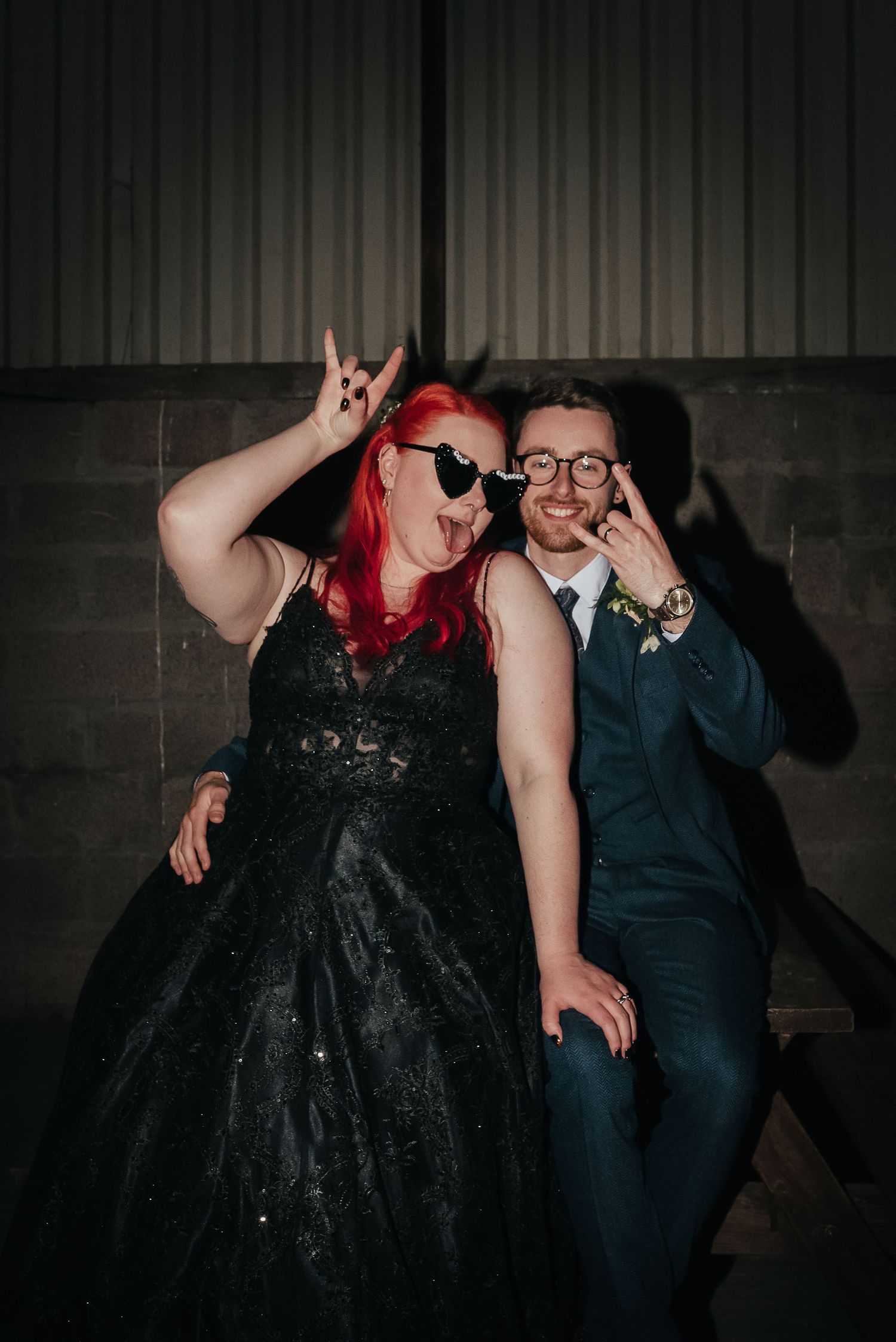 A playful couple in formal attire poses against a dark wall making peace signs.