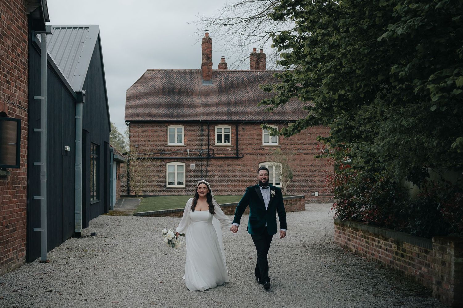 A couple walks hand in hand down a rustic path between brick buildings on their wedding day.
