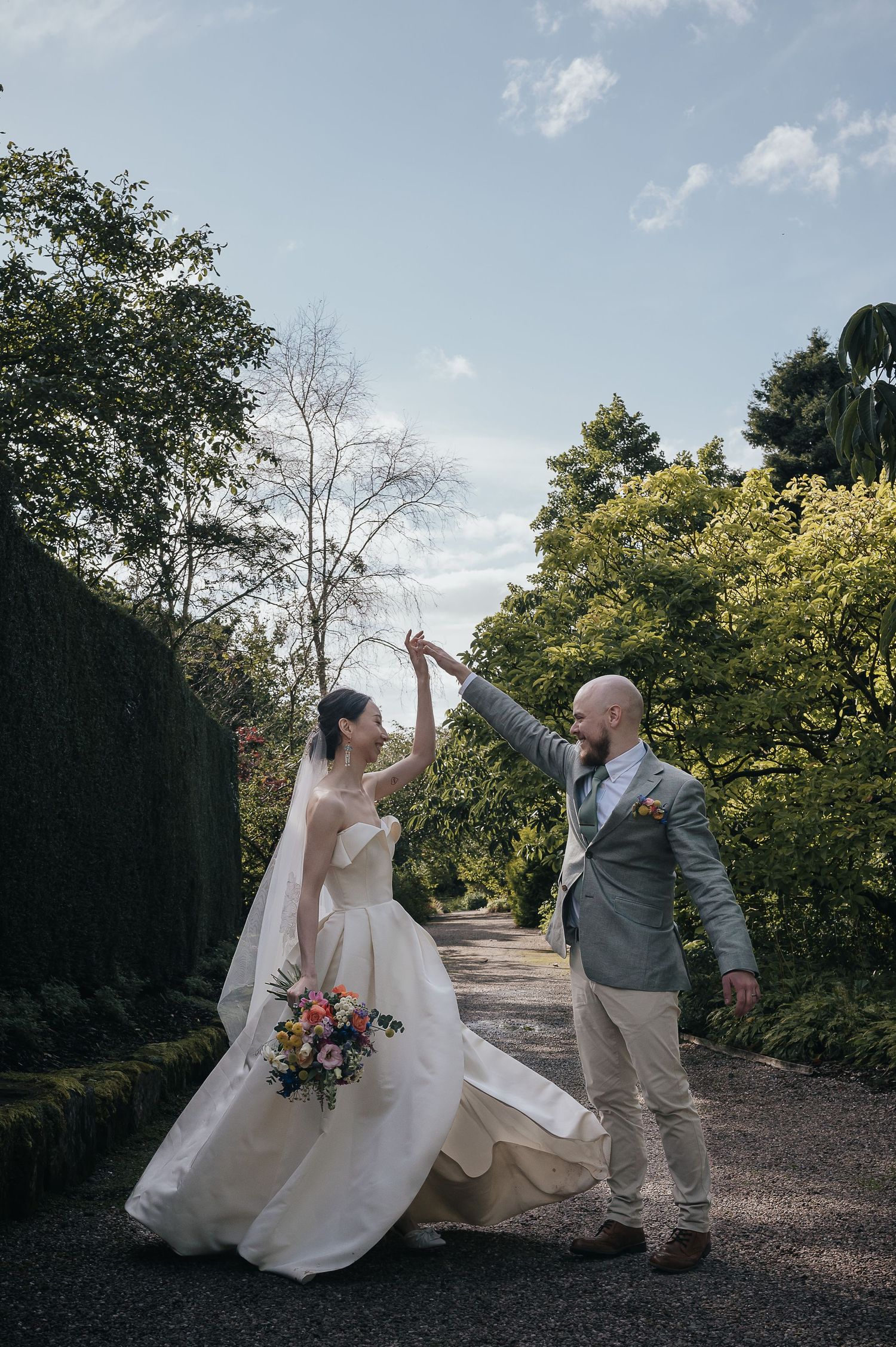 Couple dancing joyfully on garden path with bride in flowing white dress and groom in light suit.
