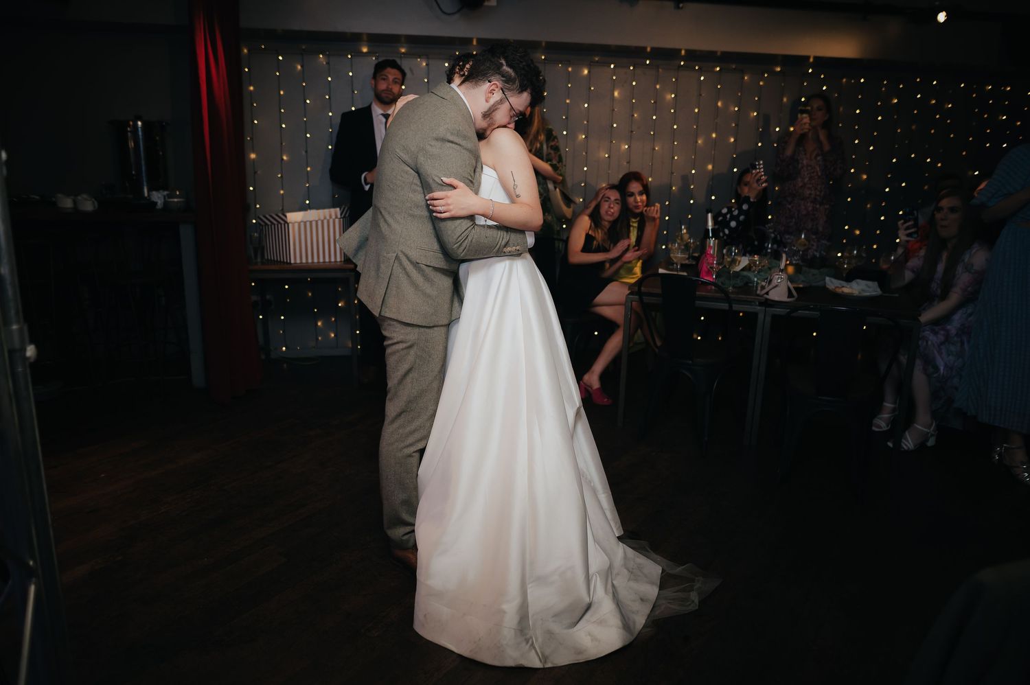 A wedding couple shares their first dance in a dimly lit reception venue with twinkle lights.