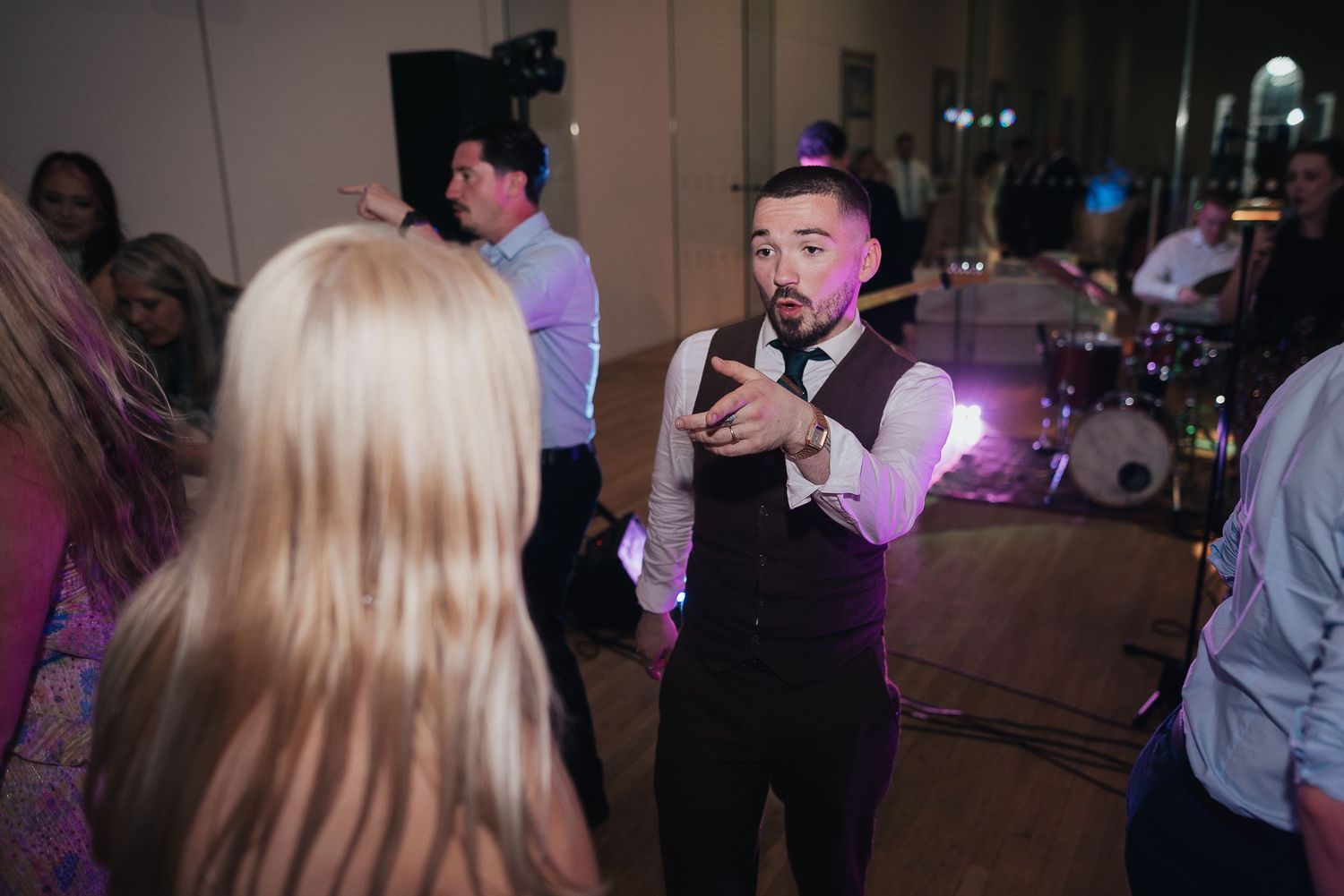 A performer in a black vest entertains guests at a wedding reception with dramatic gestures.