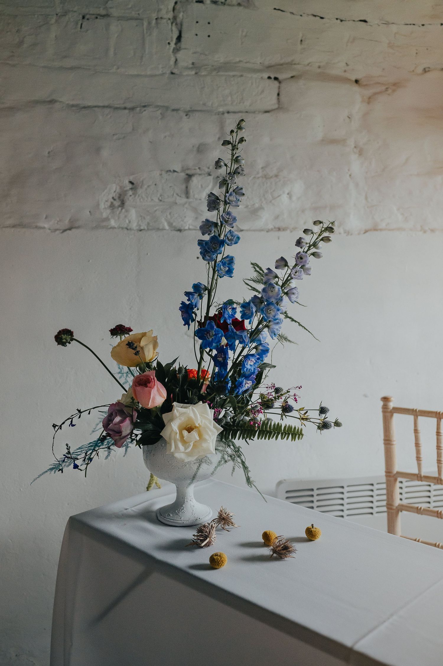 An elegant floral arrangement with blue delphiniums and roses in a white vase against a white wall.