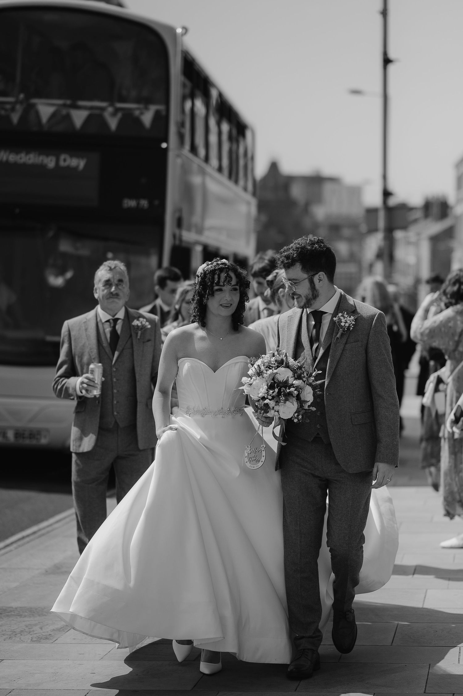 A wedding party walking on a sidewalk with a bus in the background on a sunny day.