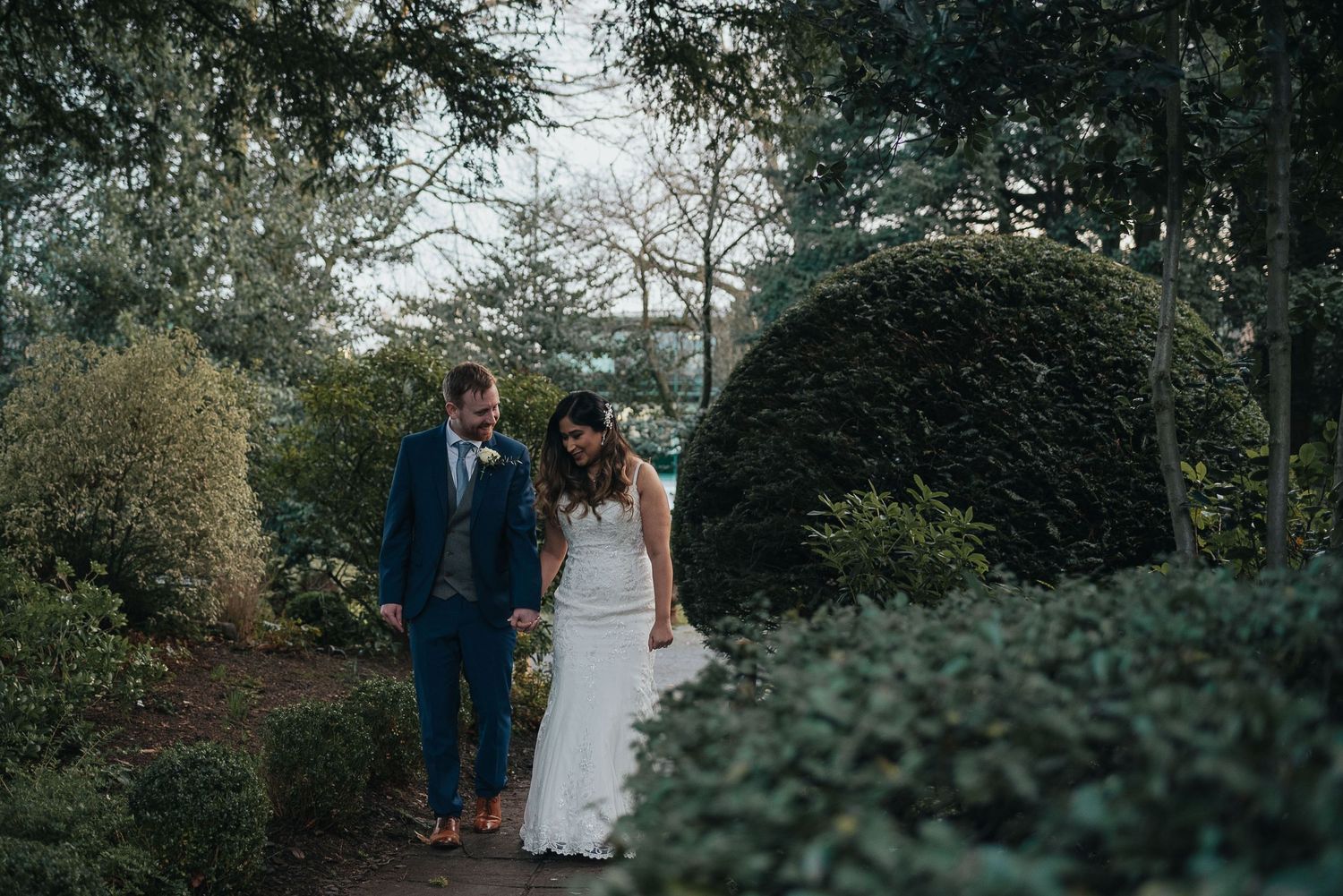 A couple walks hand in hand through a lush garden pathway with manicured hedges and greenery.
