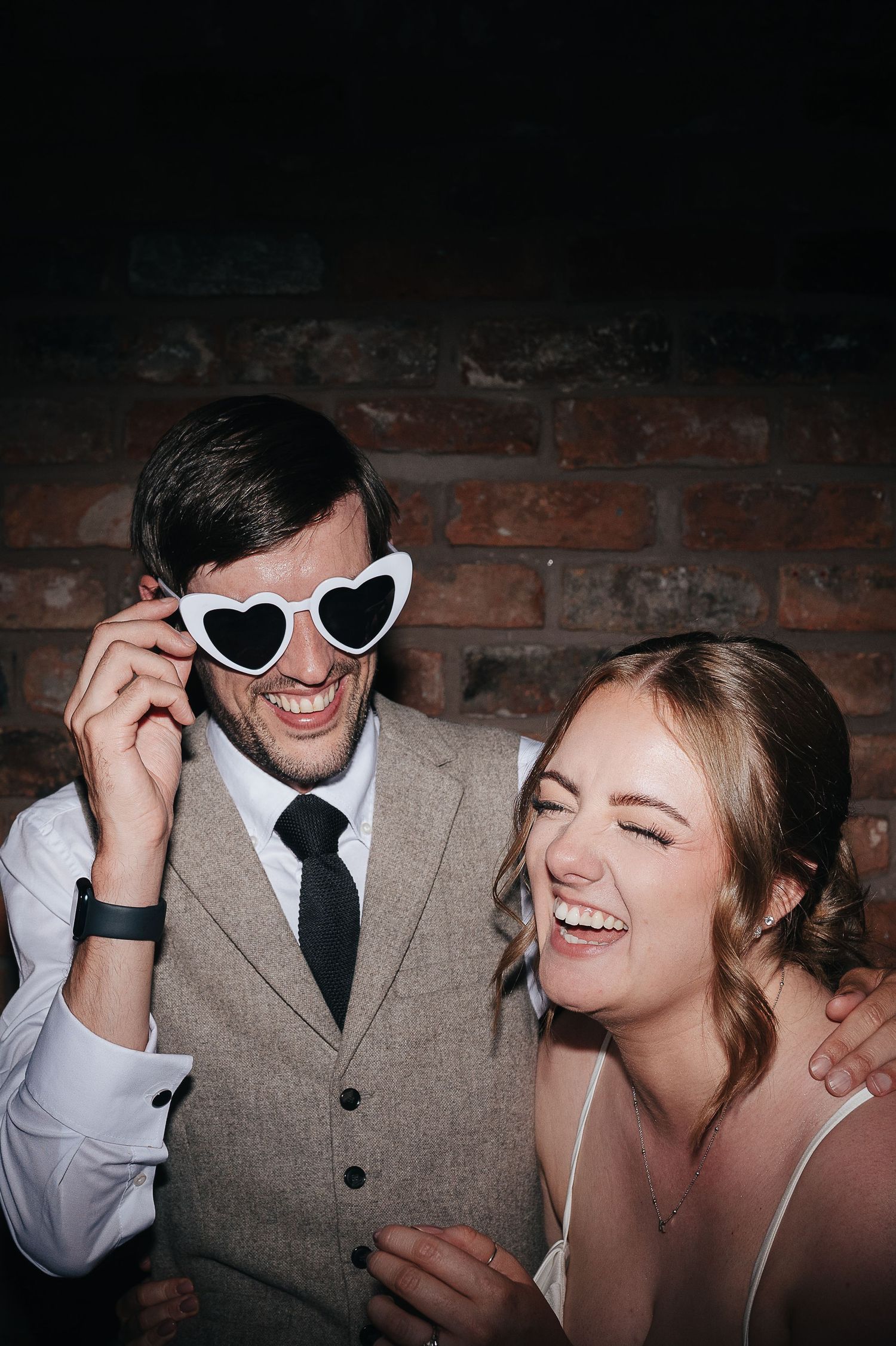 Fun wedding moment with heart-shaped sunglasses against brick wall backdrop.