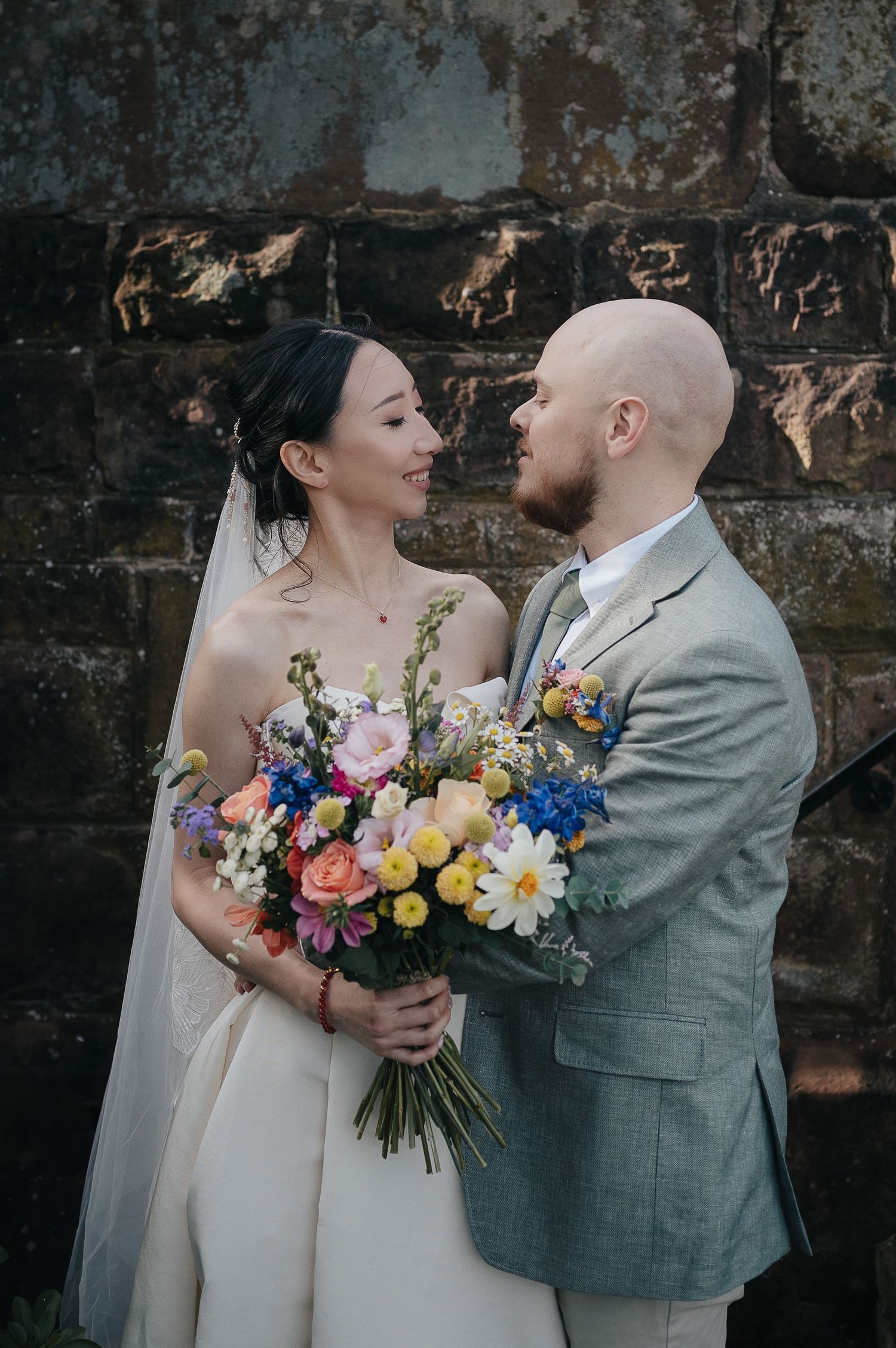A couple in wedding attire poses against a rustic wall while holding a vibrant bouquet of mixed flowers.