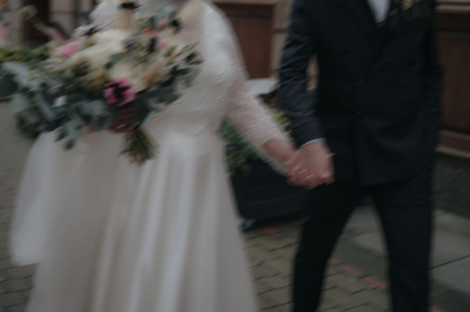 Wedding couple stands together in elegant formal attire holding floral bouquets against soft background.