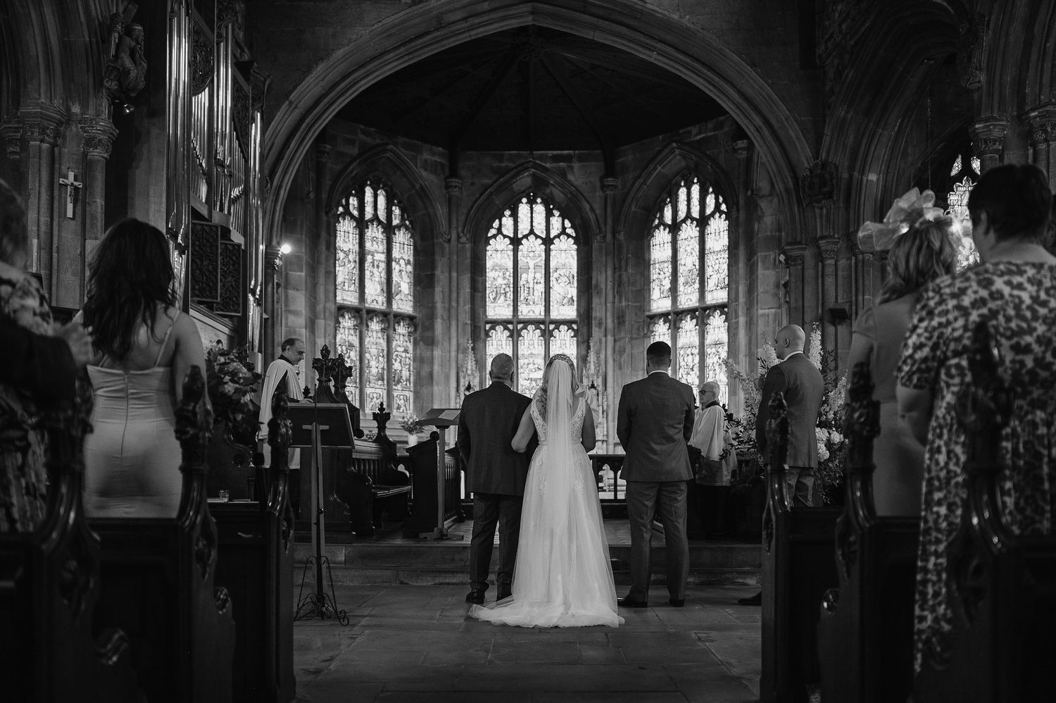 A black and white photograph of a wedding ceremony inside a beautiful gothic church with stained glass windows.