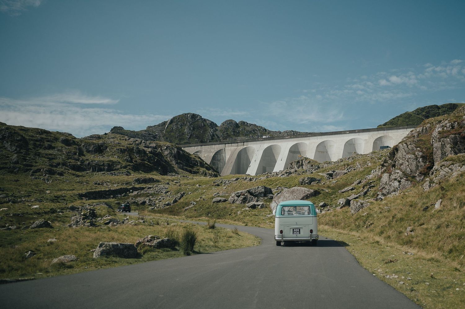 White van driving on winding mountain road past dramatic viaduct.