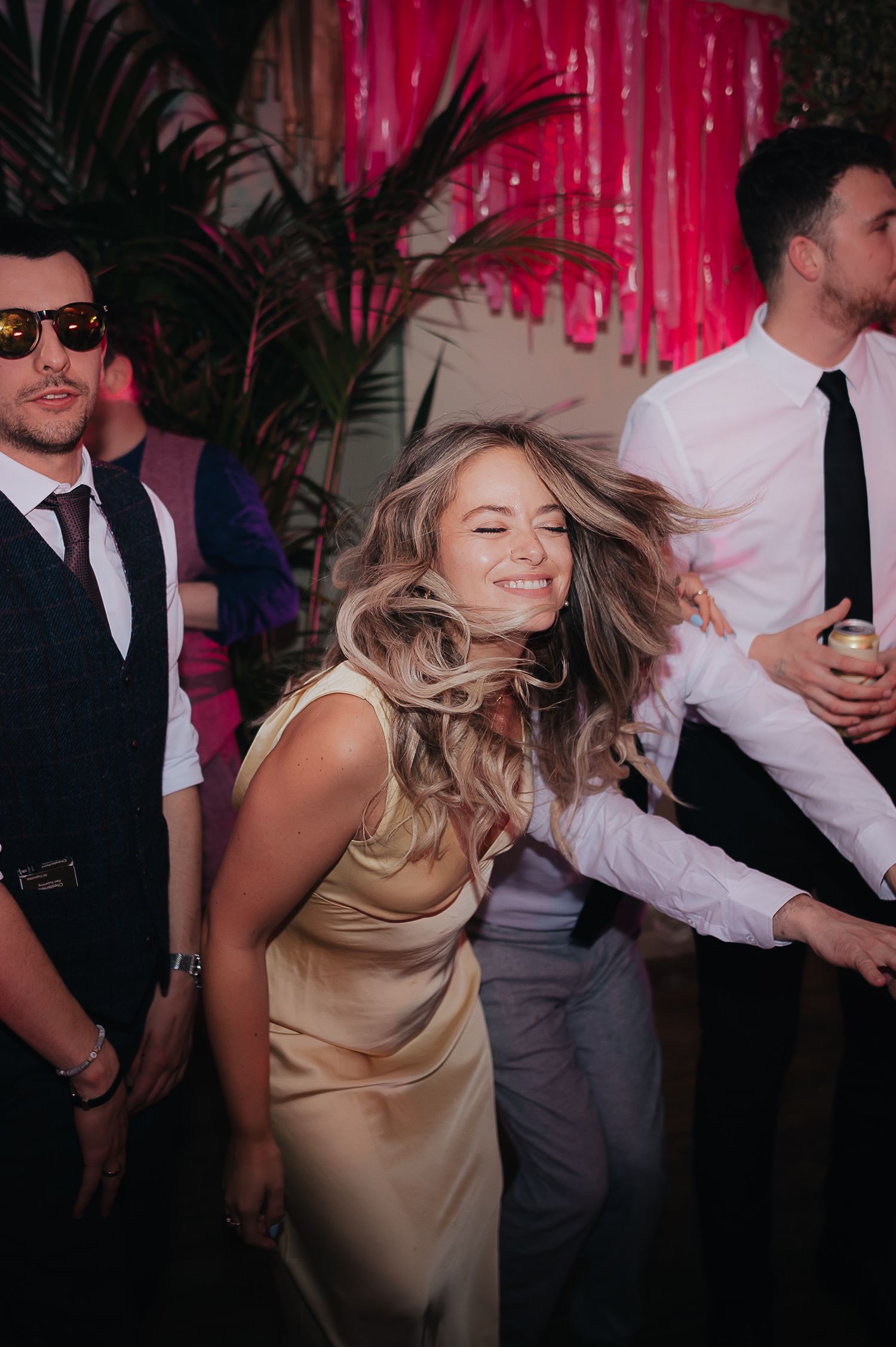 Woman in silk dress dances joyfully at a party decorated with tropical plants and pink streamers.